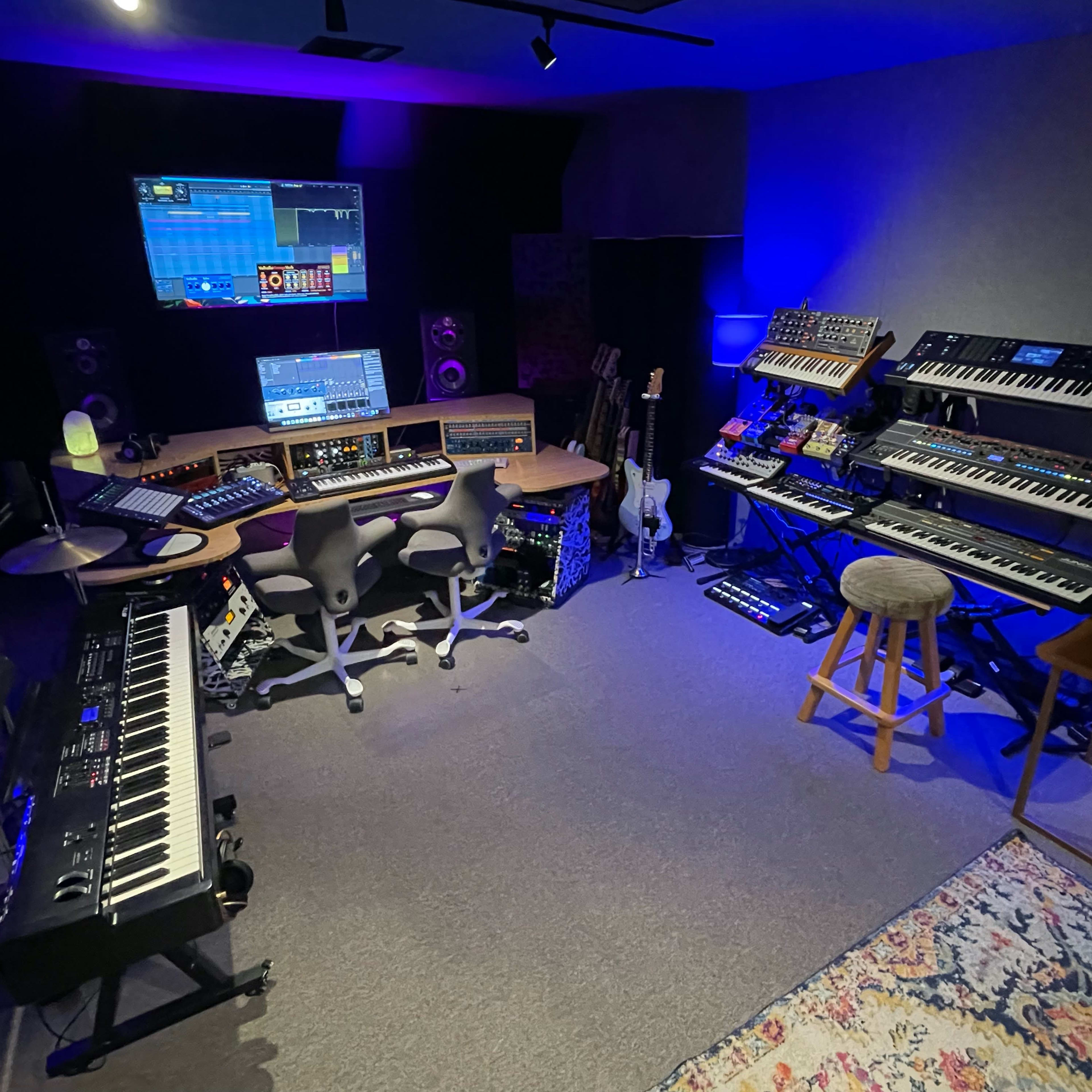 A music studio filled with various synthesizers, keyboards, and musical equipment arranged around a central workstation.