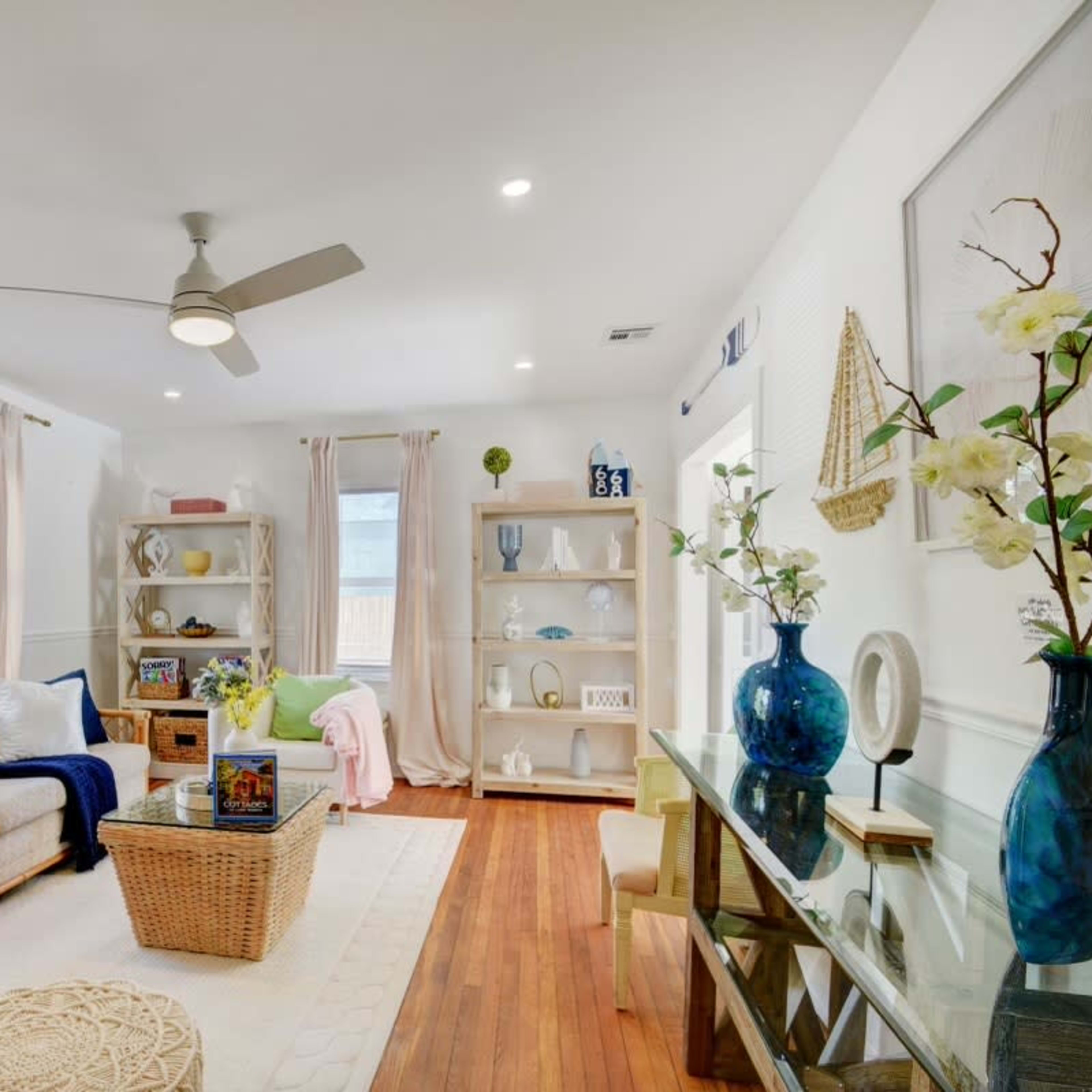 A bright living room featuring a sofa, a coffee table, and decorative shelves filled with plants and books, complemented by a ceiling fan and large windows.