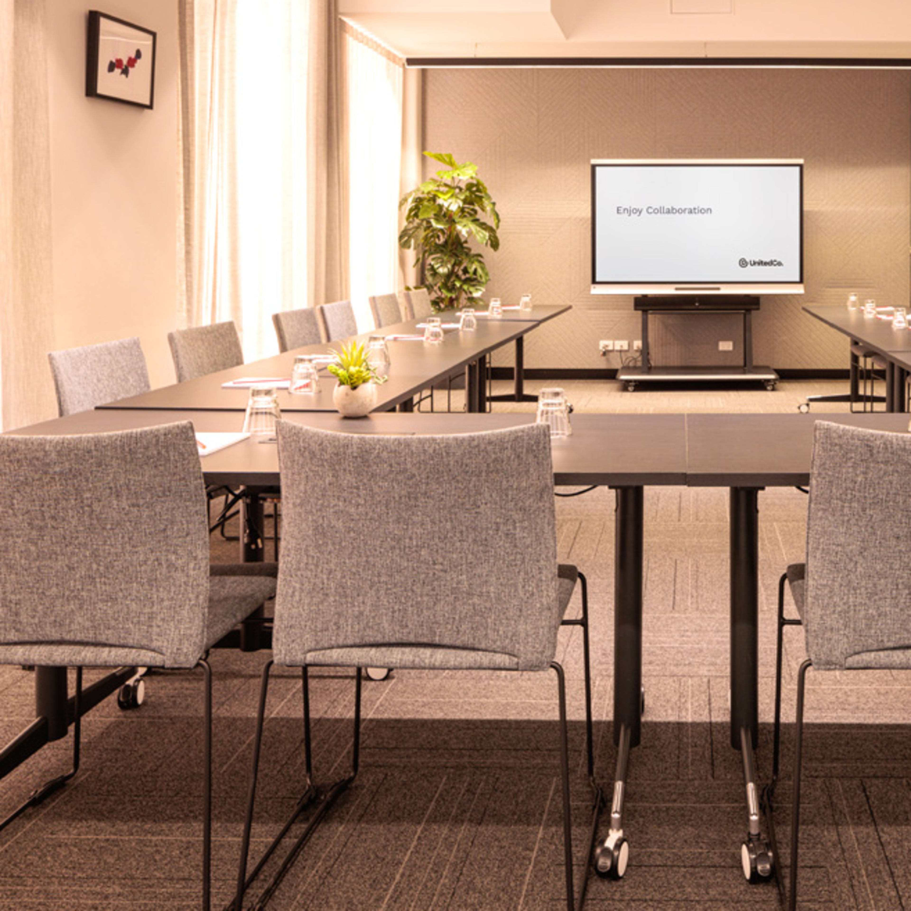 A modern conference room is set up with multiple rectangular tables arranged in a U-shape, each with chairs and water bottles, and features a large screen displaying "Enjoy Collaboration."