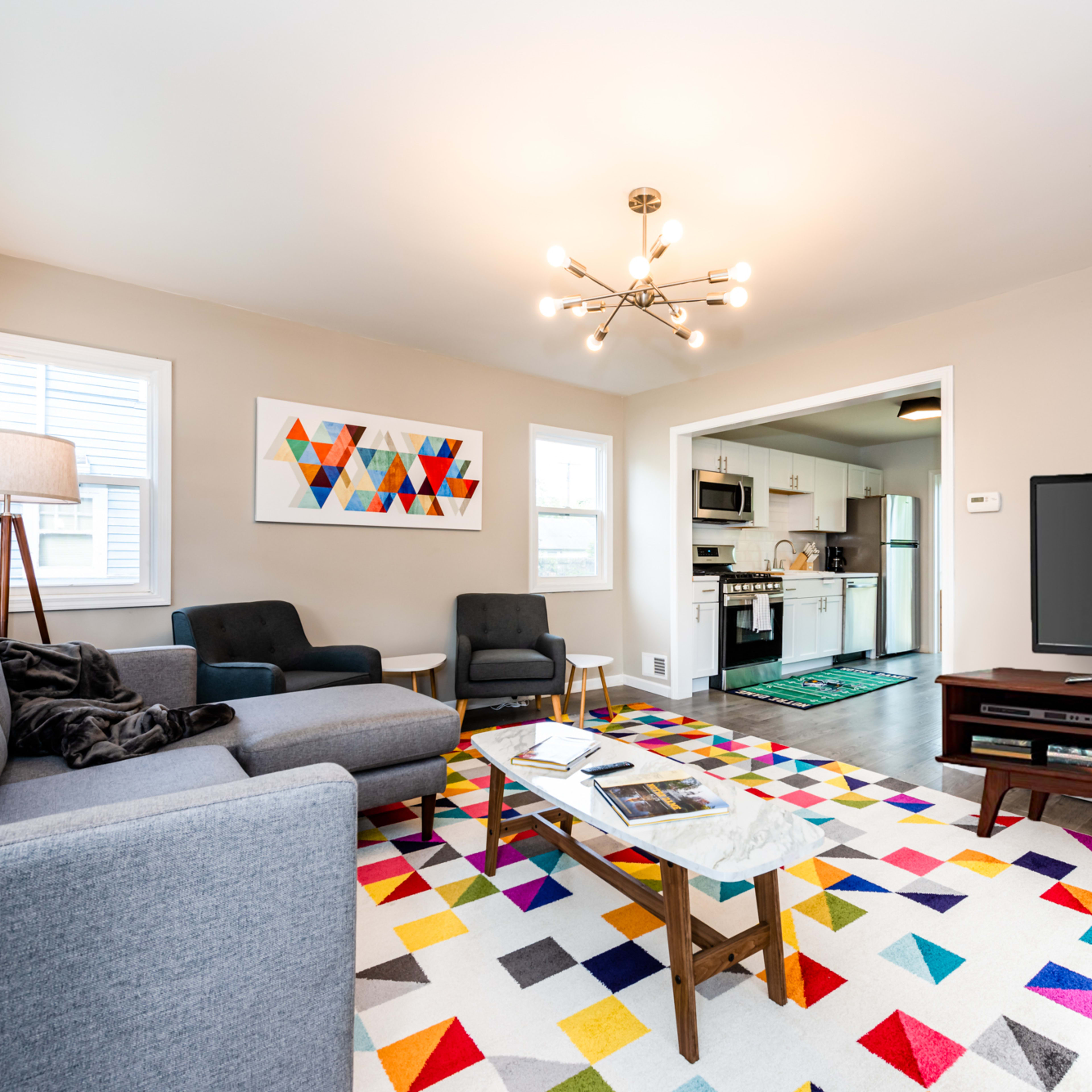 A modern living room features a gray sofa, colorful patterned rug, and a television cabinet, with an adjacent kitchen visible in the background.