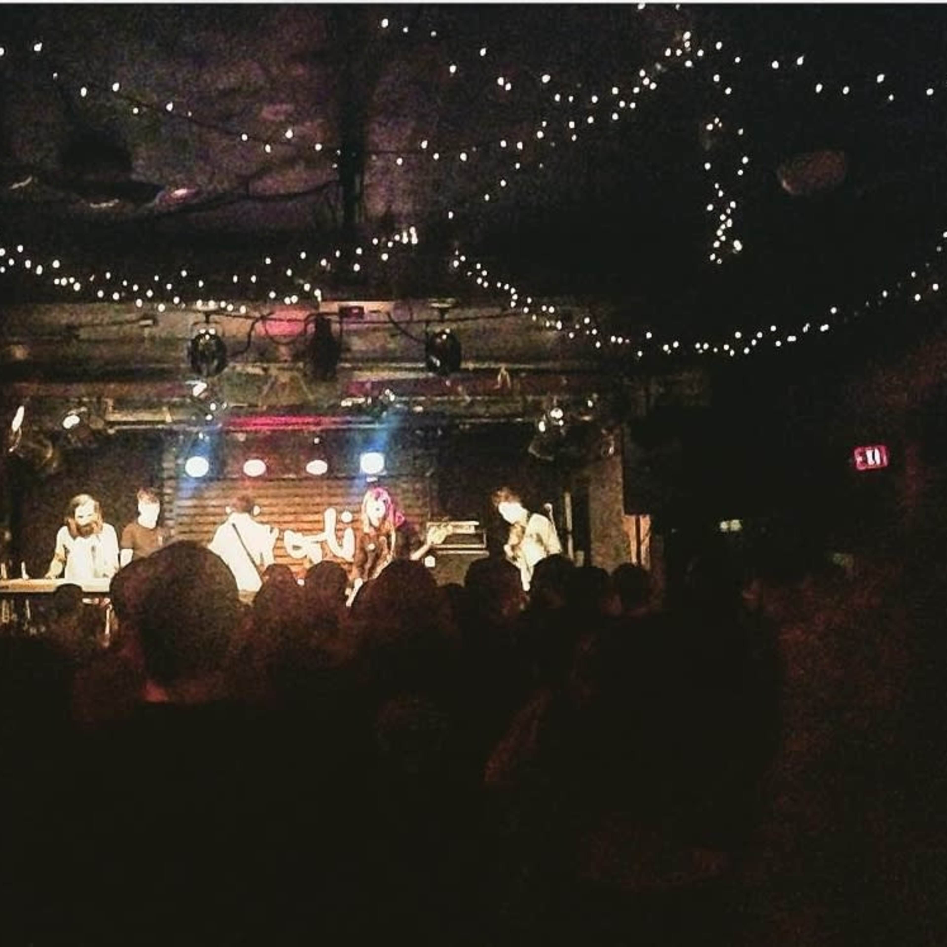 A band performs on stage in a dimly lit venue filled with an audience, decorated with string lights overhead.