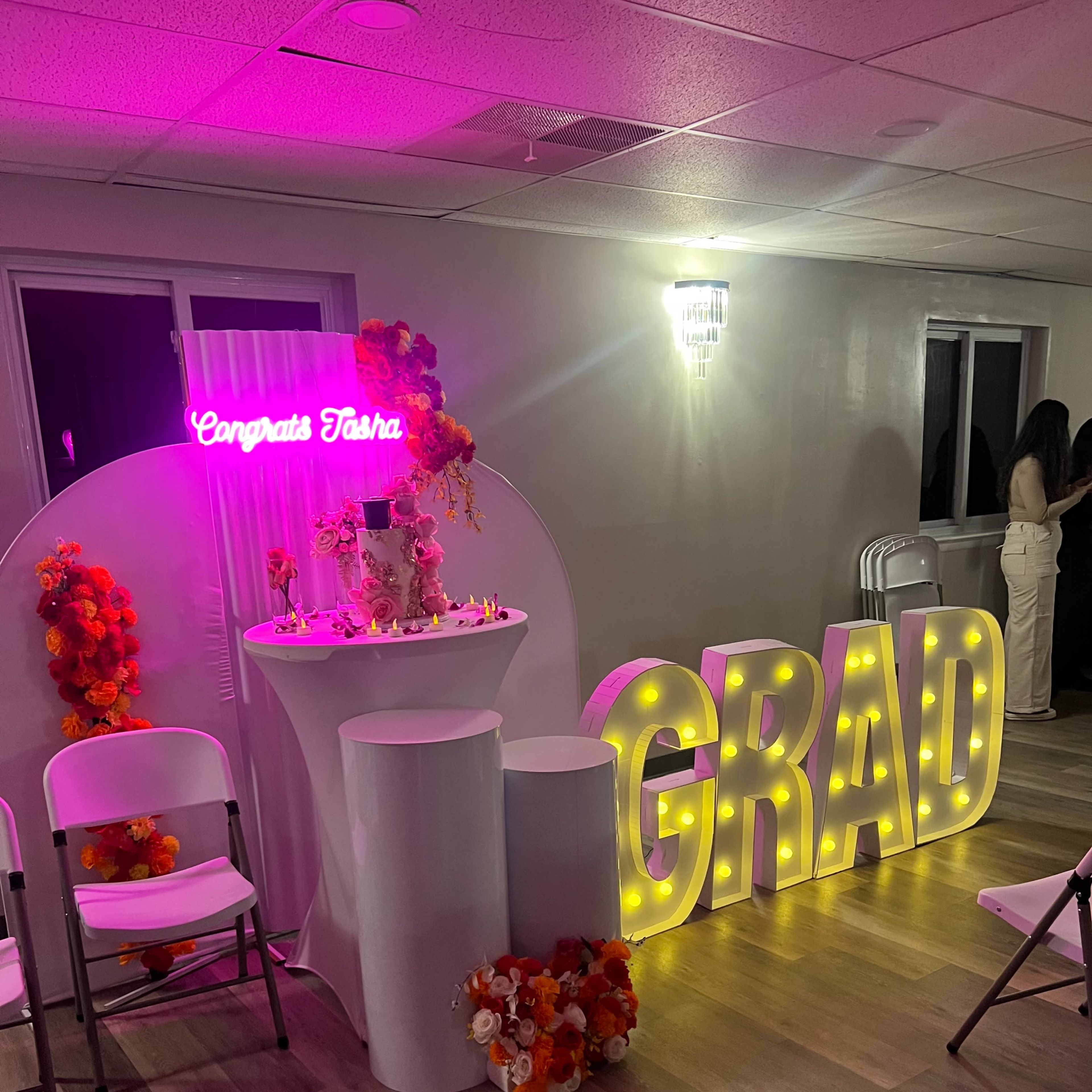 A decorated area for a graduation celebration featuring a lit "GRAD" sign, a table with floral arrangements, and a neon sign that reads "Congrats Tasha."