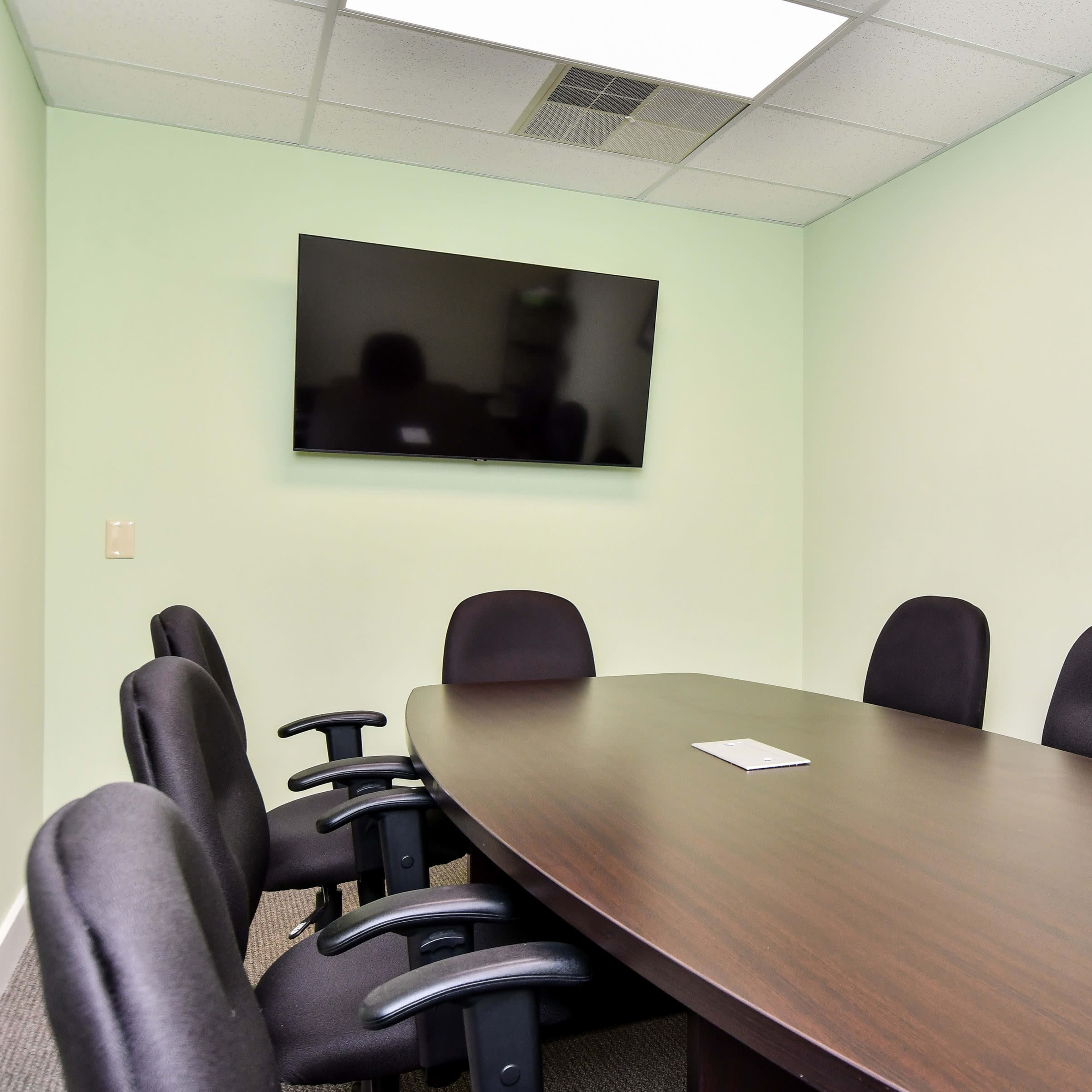 A conference room features a large wooden table surrounded by six black chairs, with a mounted television and a framed photo on the green wall.