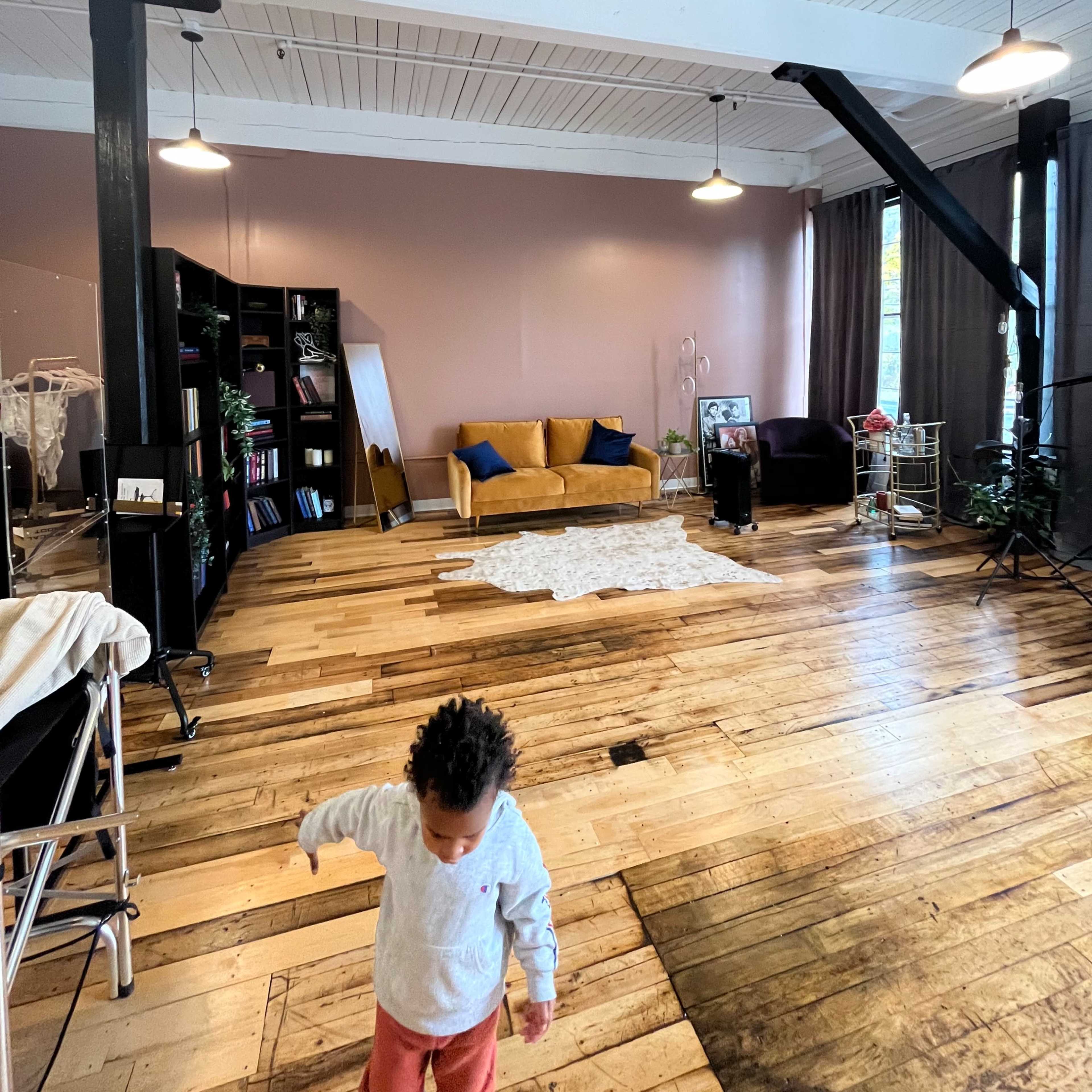 A child walks across a spacious room with wooden floors, featuring a couch, a bookshelf, and large windows.