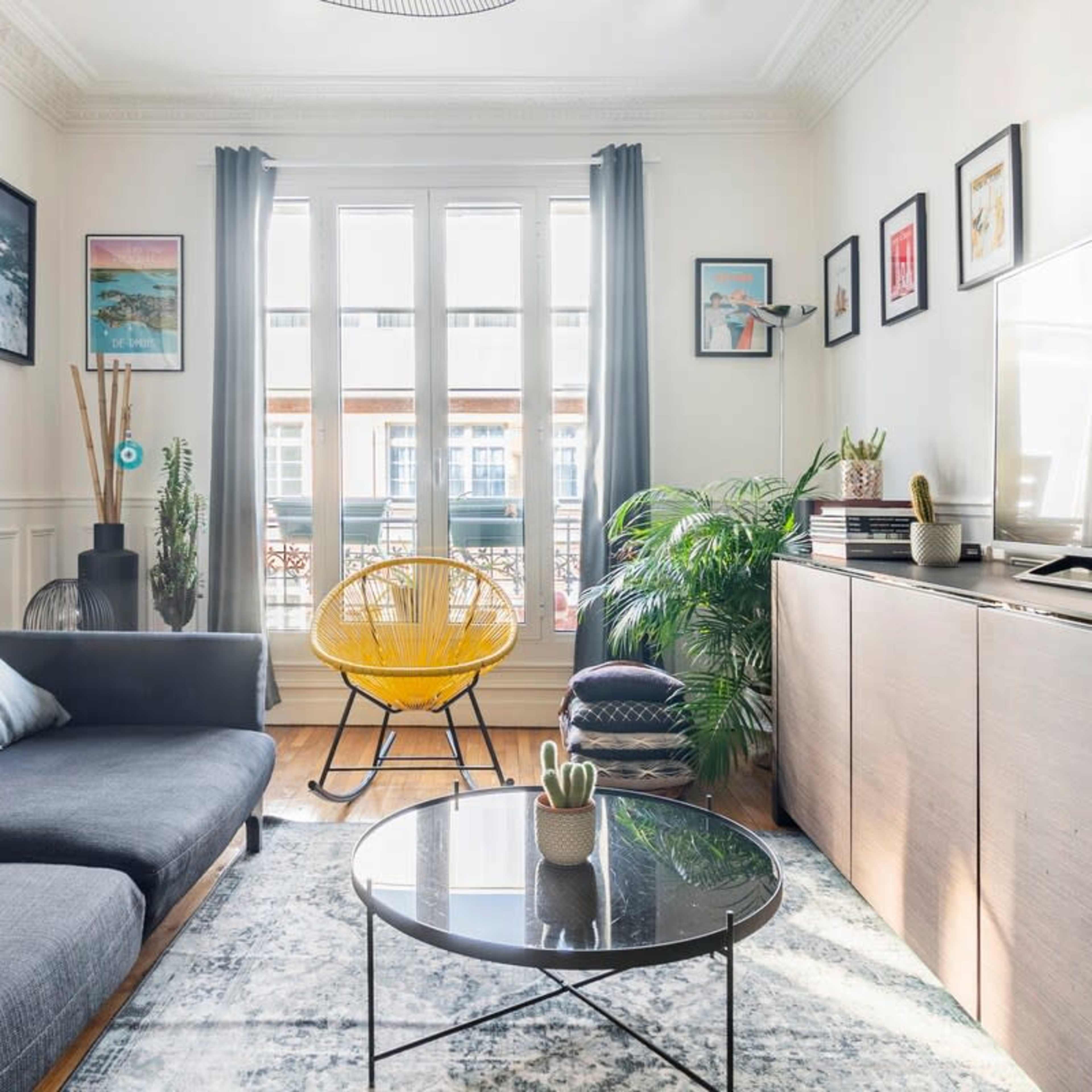 A modern living room features a dark sofa, a yellow chair, a round black coffee table, and various plants, with framed artwork hanging on the walls.