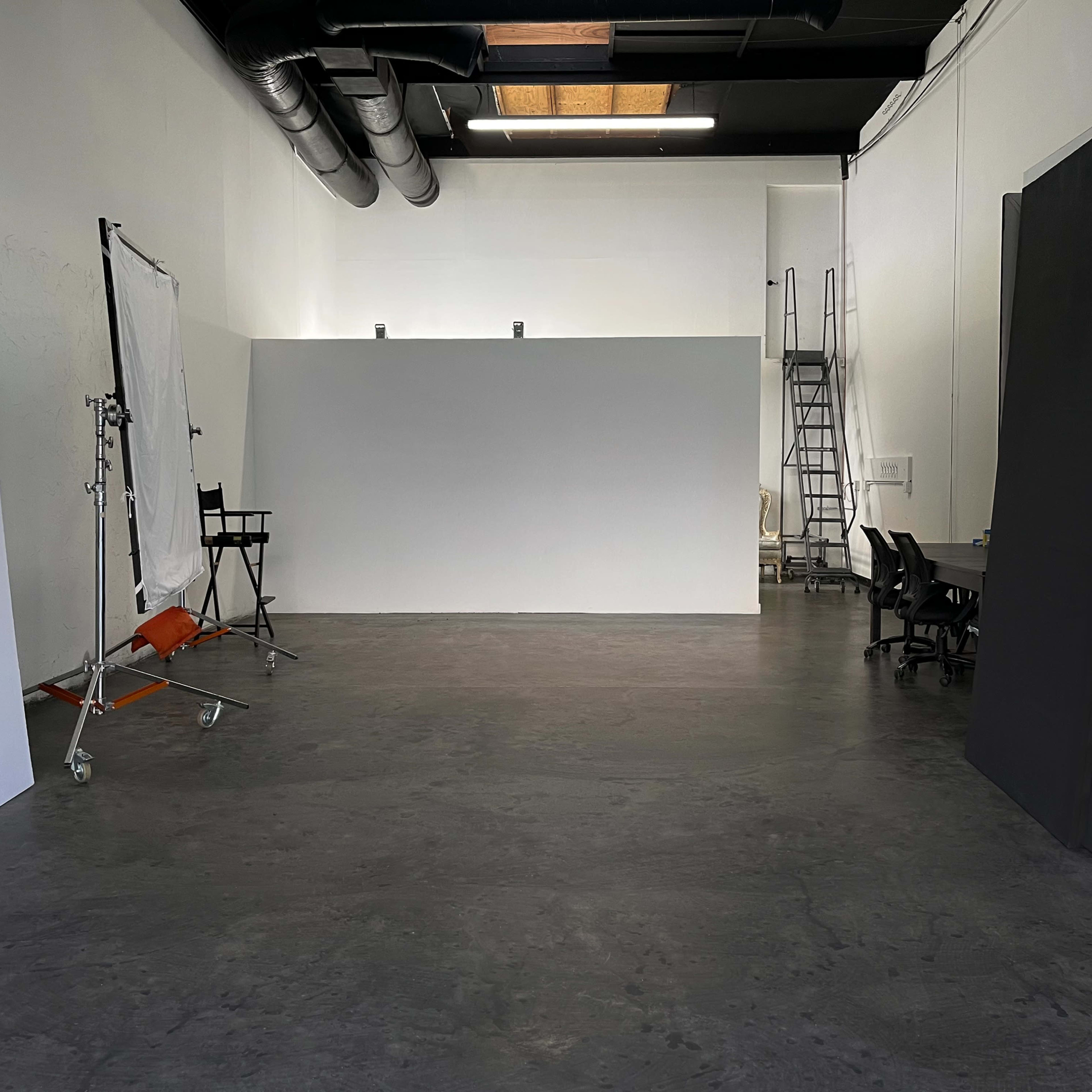 A spacious studio with a large empty backdrop, a few chairs, and a ladder against the wall.