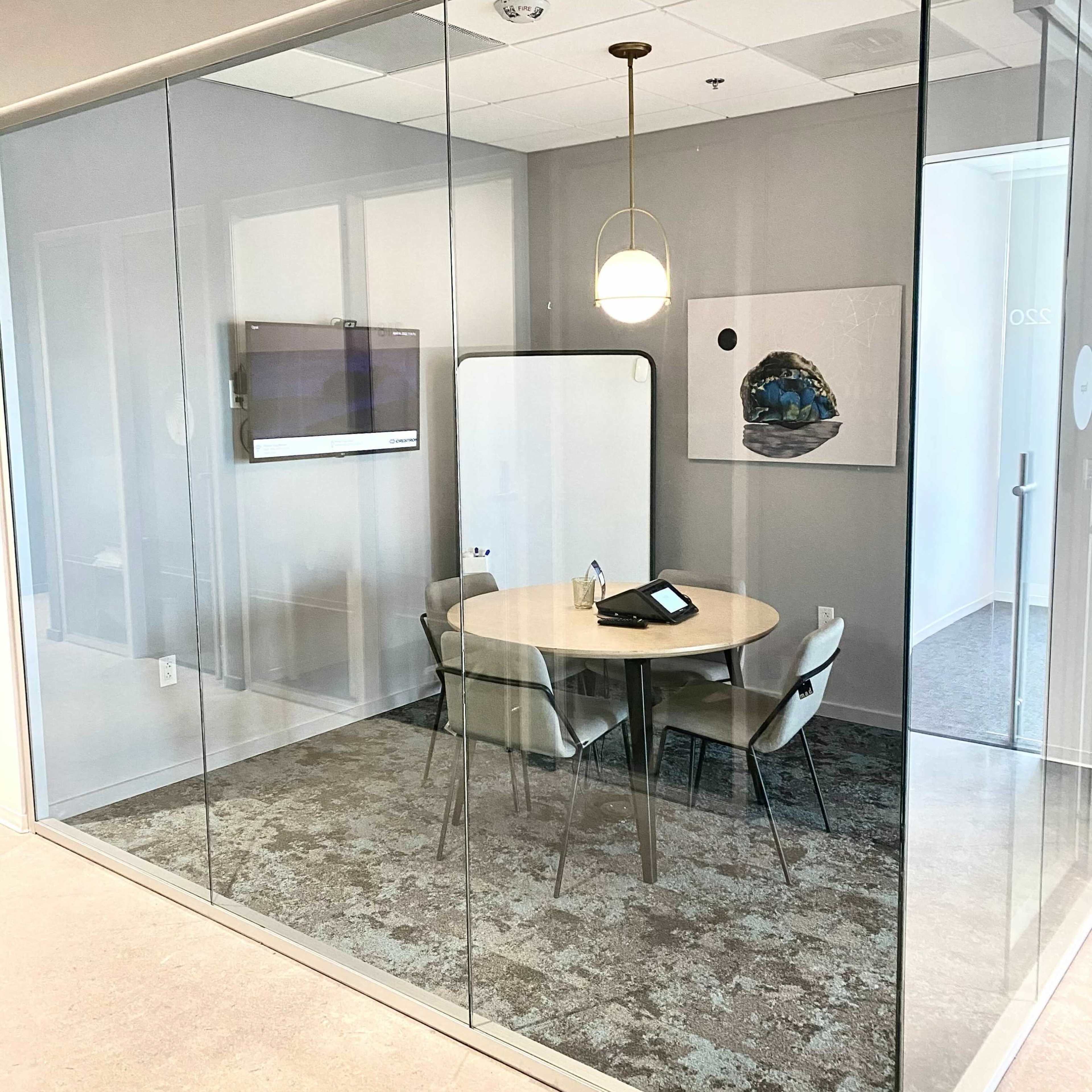 A modern conference room enclosed by glass walls, featuring a round table surrounded by chairs and a presentation screen on one wall.