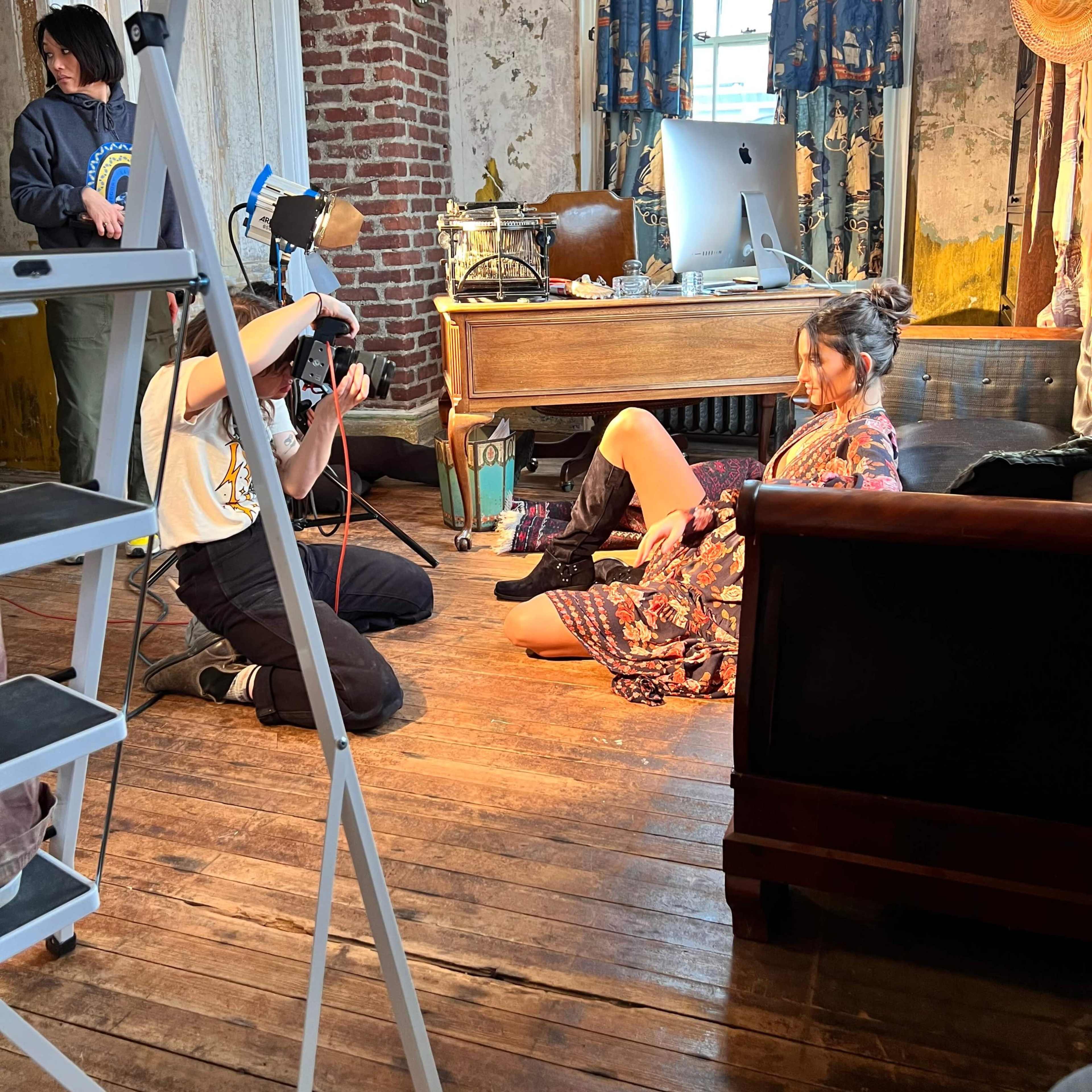 A photographer captures an image of a model sitting on the floor in a vintage-styled room while another person assists in the background.