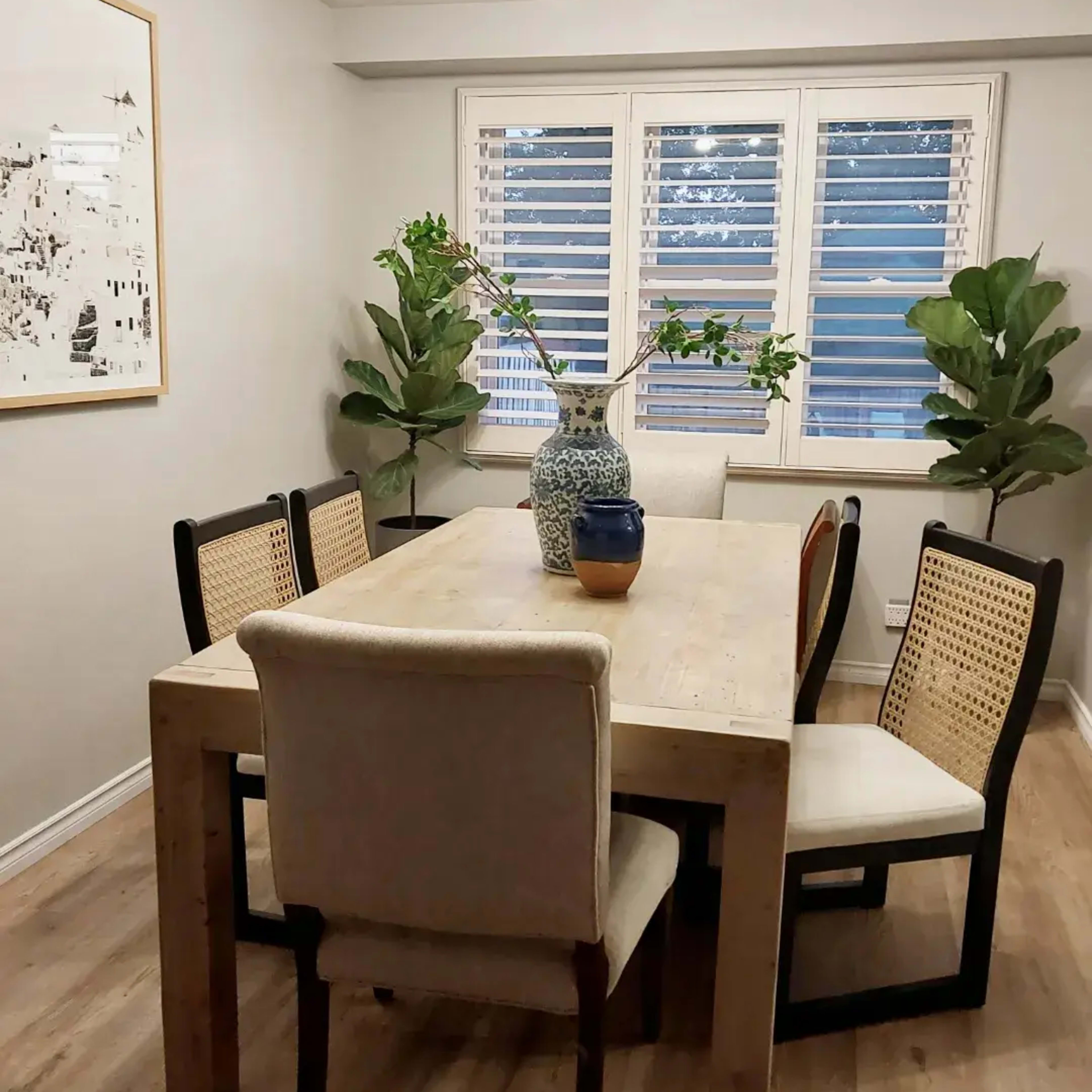 A dining room features a wooden table surrounded by six chairs, with large plants and a vase on the table, beneath a modern light fixture.
