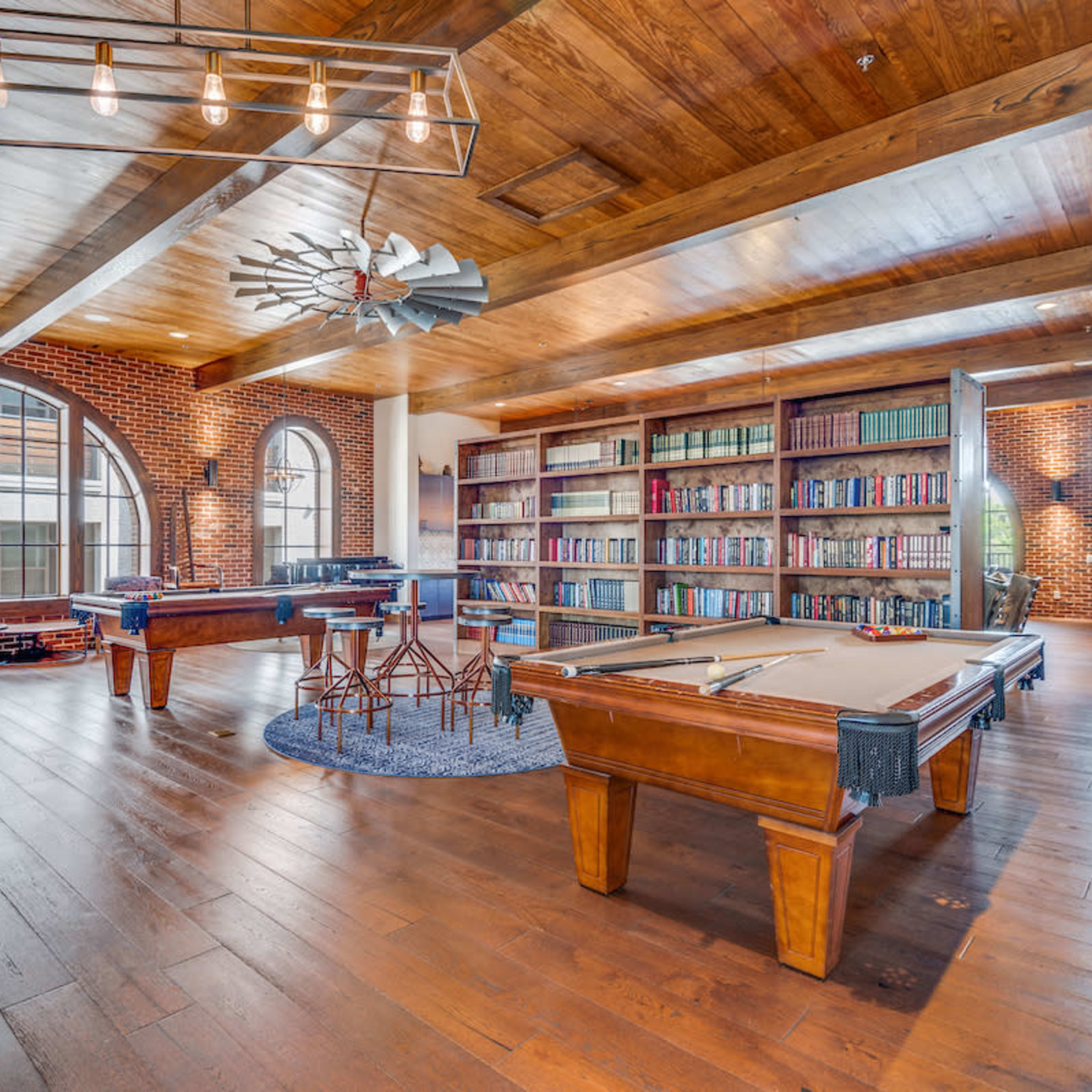 The scene features a spacious room with exposed brick walls, wooden beams, a pool table, and bookshelves filled with books.