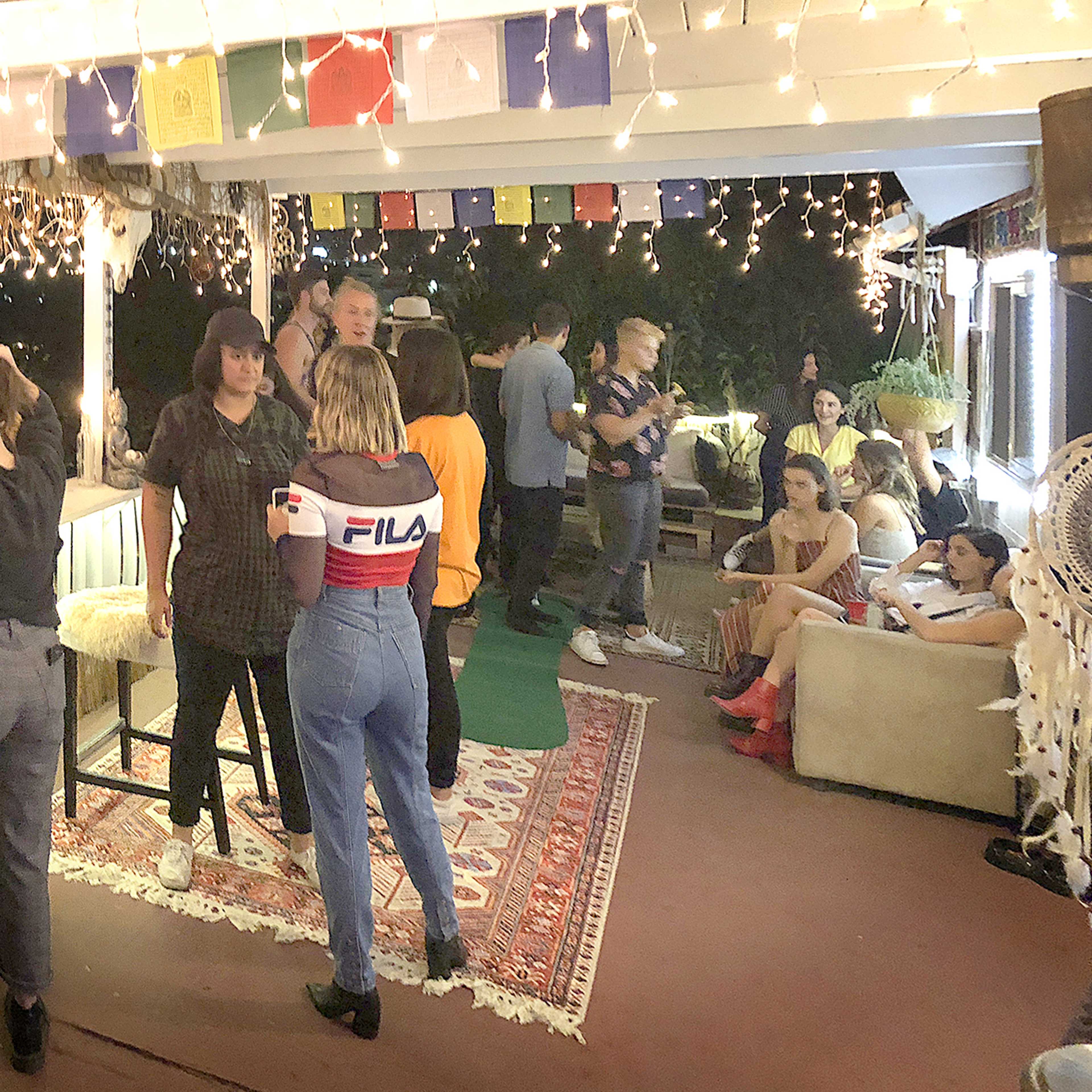A group of people socializes on a covered porch decorated with string lights and colorful flags.