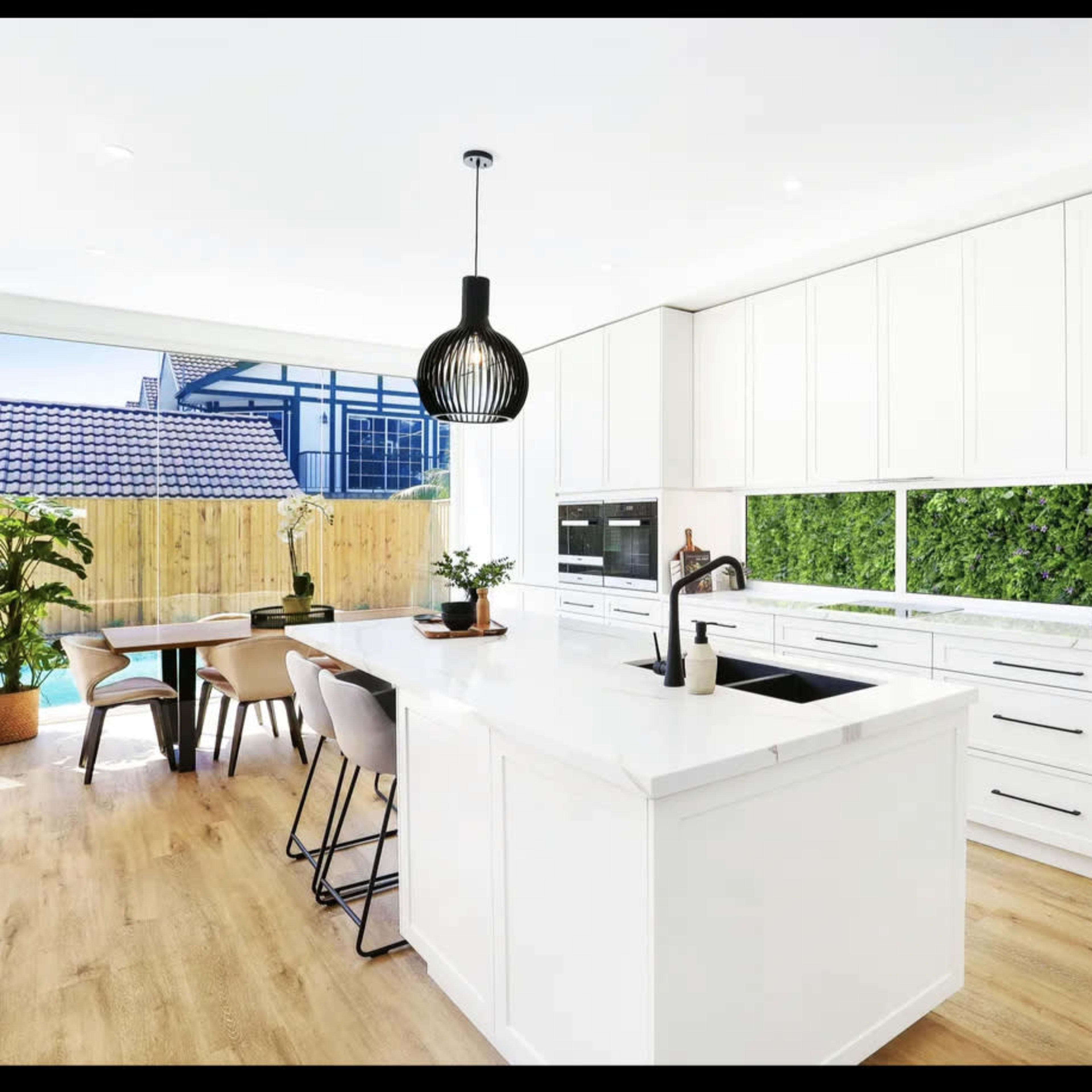 A modern kitchen features a central island with a sink, surrounded by sleek cabinetry and natural greenery visible through large windows.