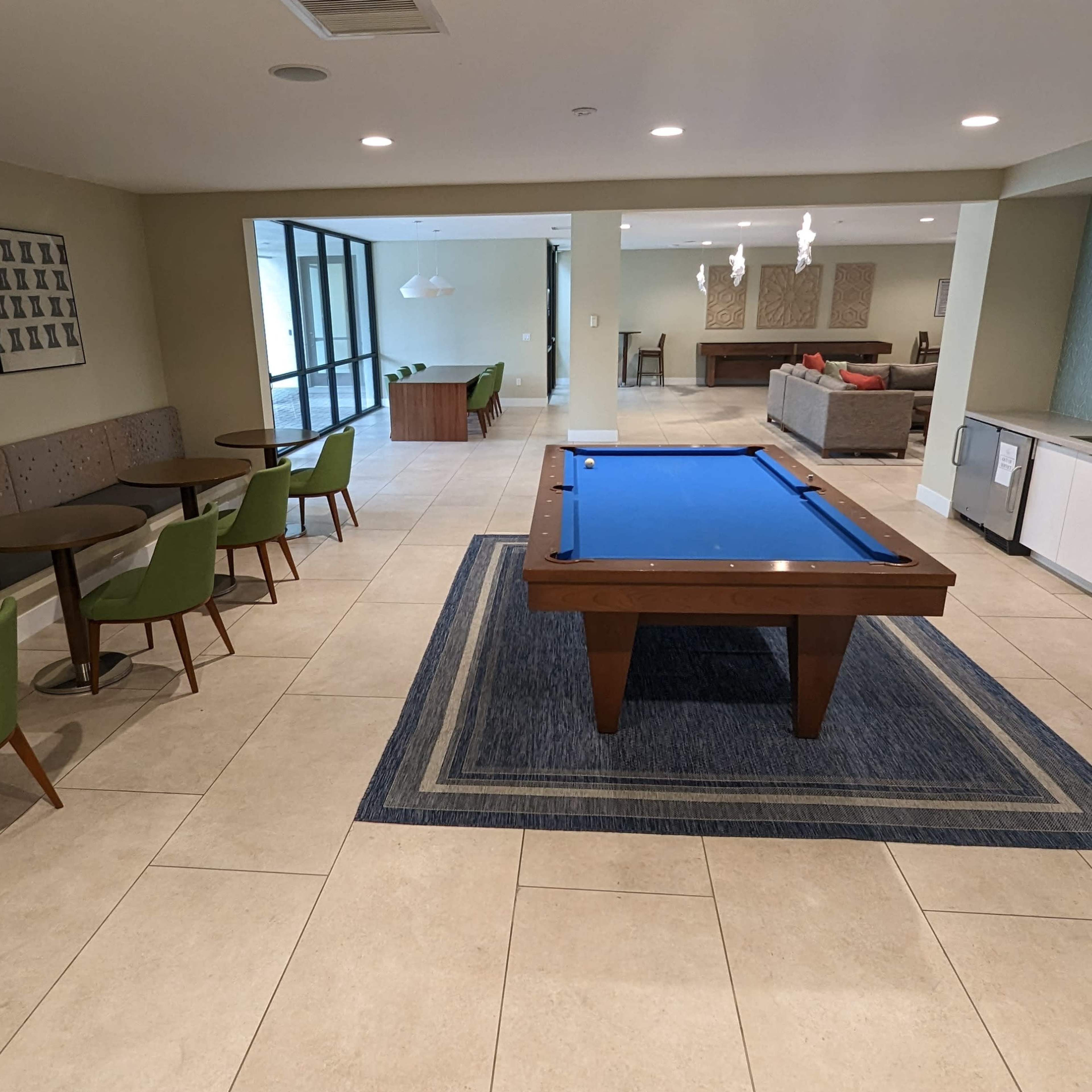 A spacious lounge area featuring a pool table, seating with small tables, and a kitchenette along one wall.