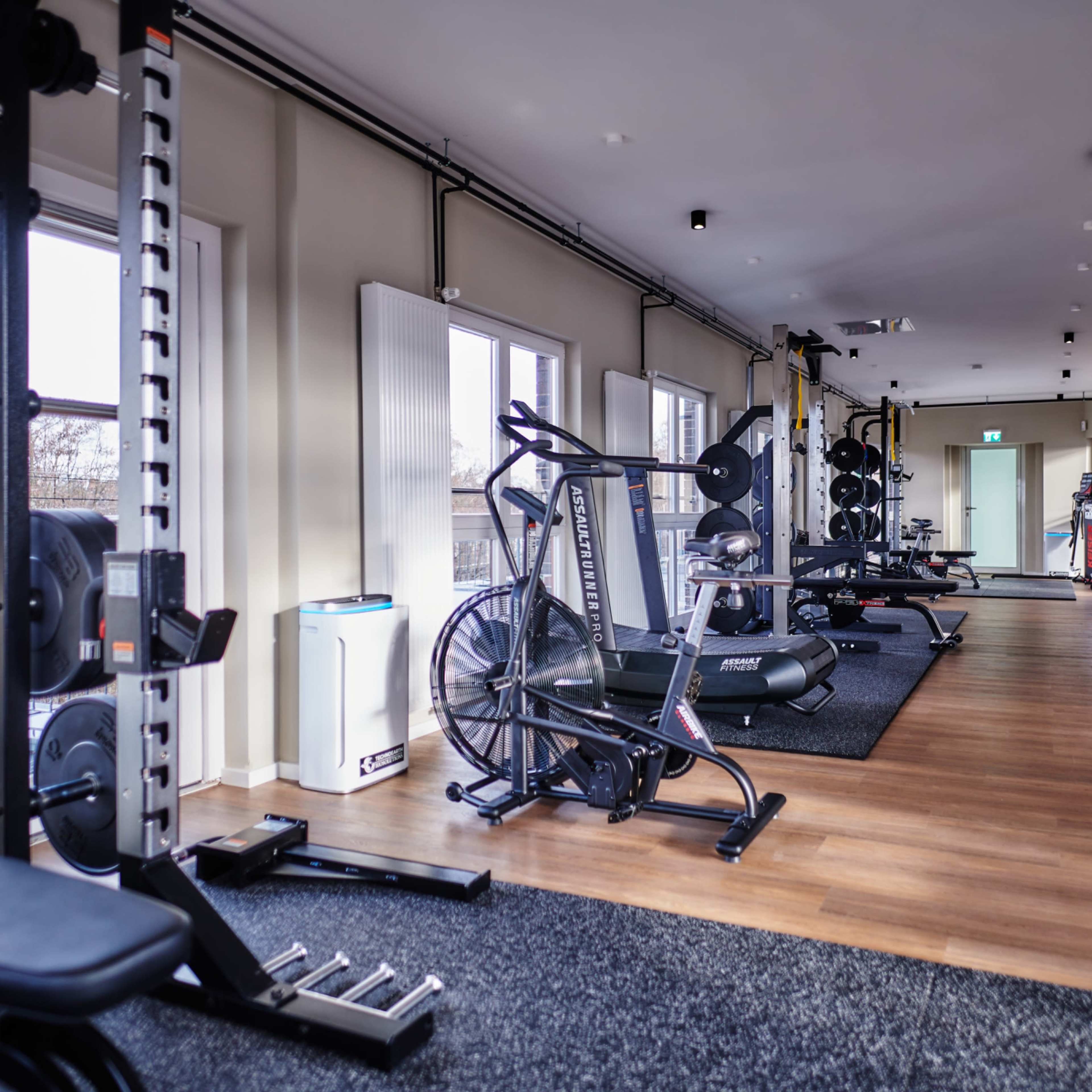 A modern gym interior with various fitness equipment arranged along a long, well-lit room featuring windows and wooden flooring.