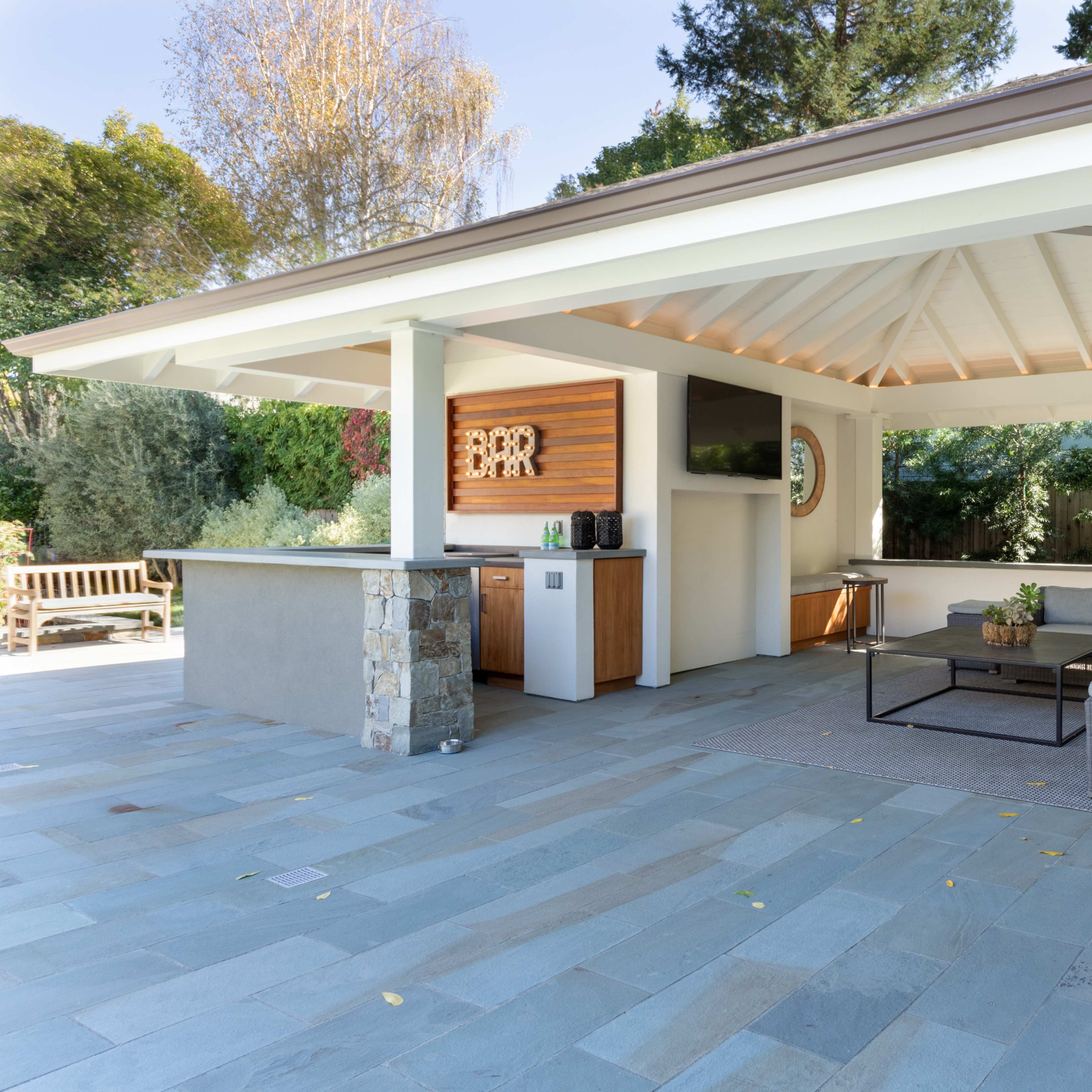 A covered outdoor seating area with a stone bar, gray sectional sofa, and a large flat-screen TV in a landscaped backyard.