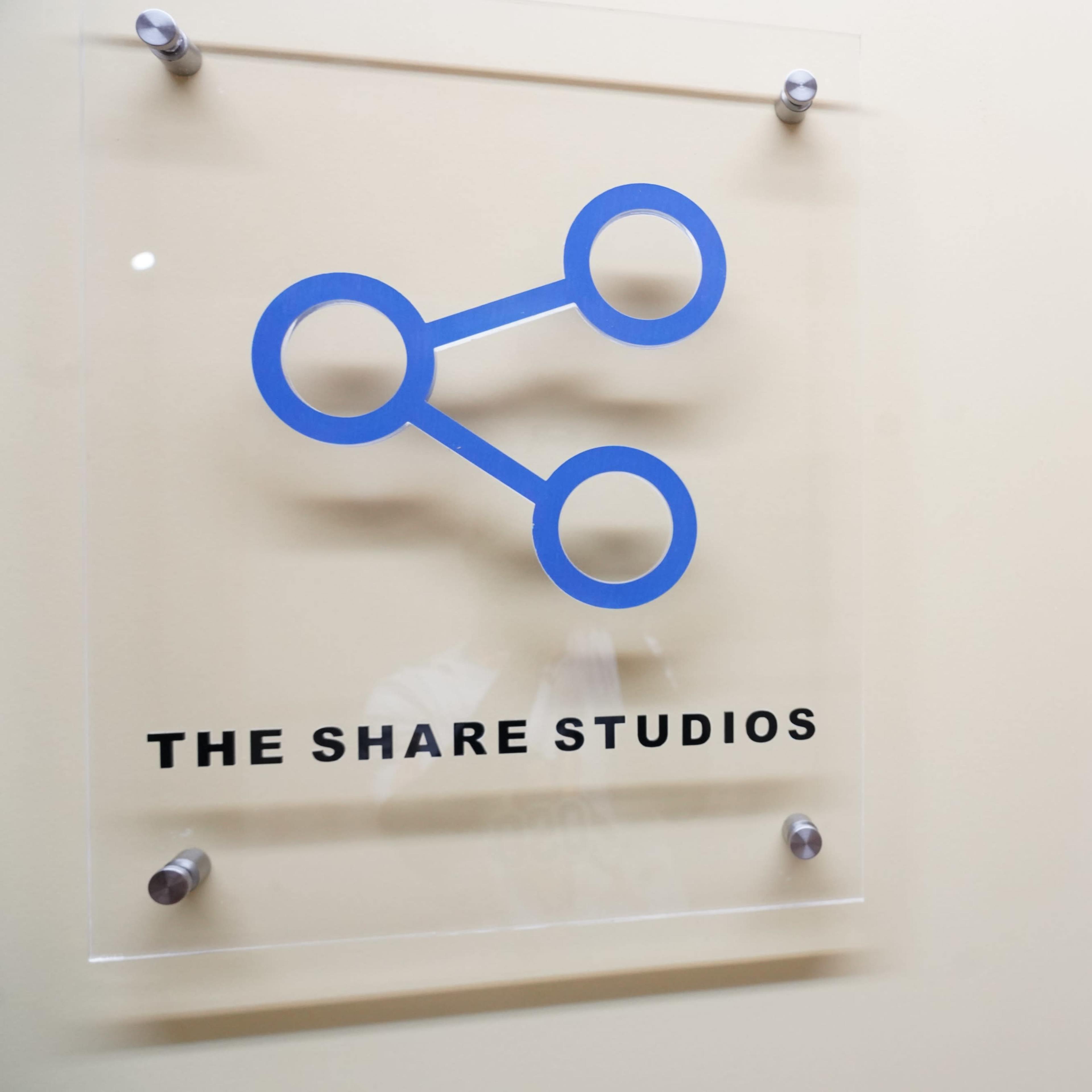 A sign displaying the logo of "The Share Studios" is mounted on a wall near an office interior with framed images visible in the background.