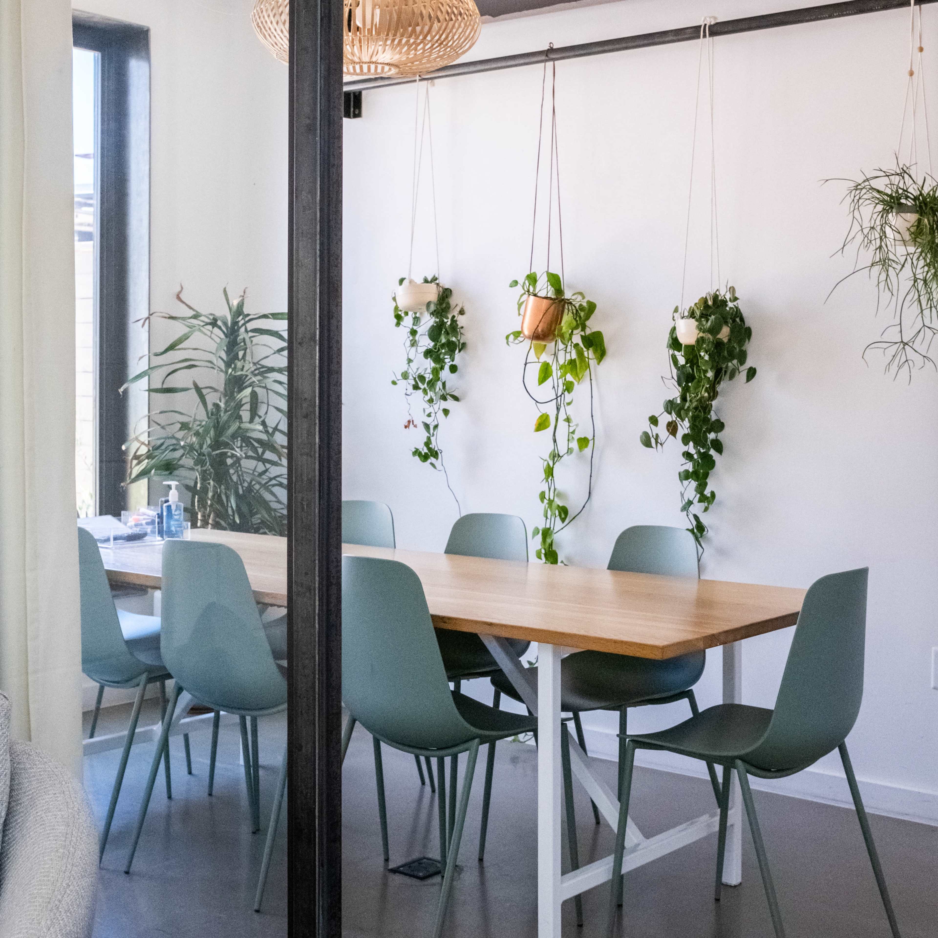 A modern dining area features a wooden table surrounded by green chairs, with hanging plant pots and a large window providing natural light.