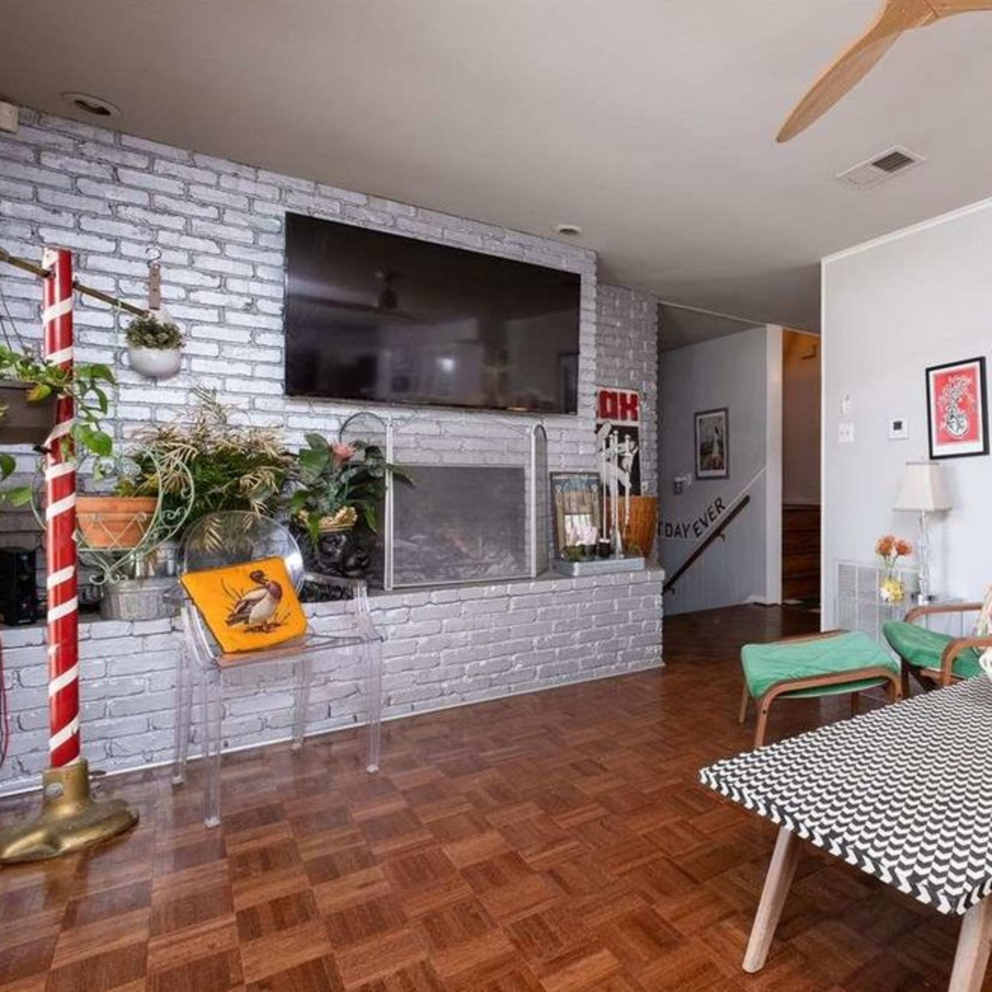 The living room features a brick fireplace surrounded by potted plants, a television mounted above, and seating arranged around a patterned coffee table.
