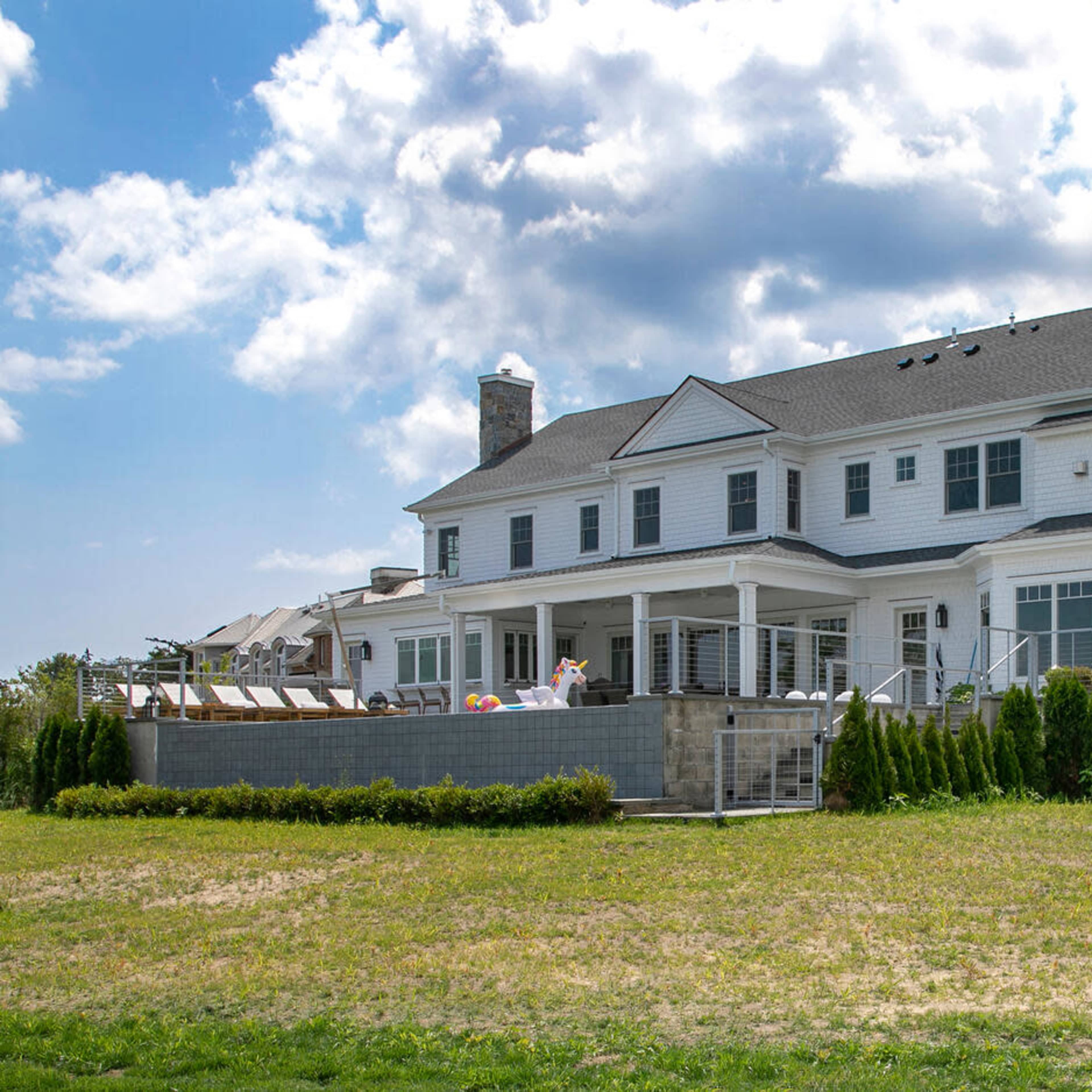 A large, two-story white house with a stone chimney features a spacious deck and a manicured lawn.