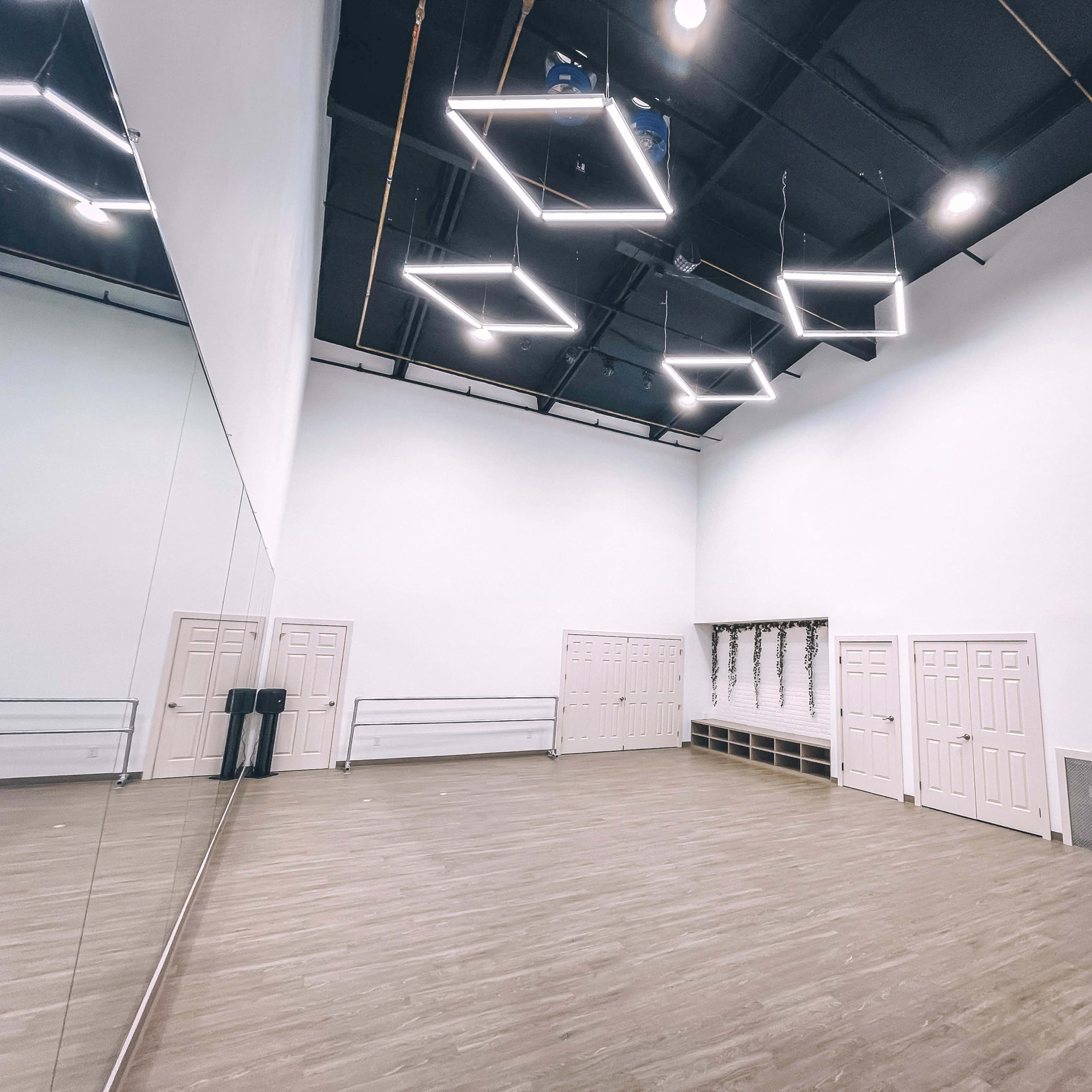 A spacious, empty studio with wooden floors, a large wall mirror, and several doors along one side.