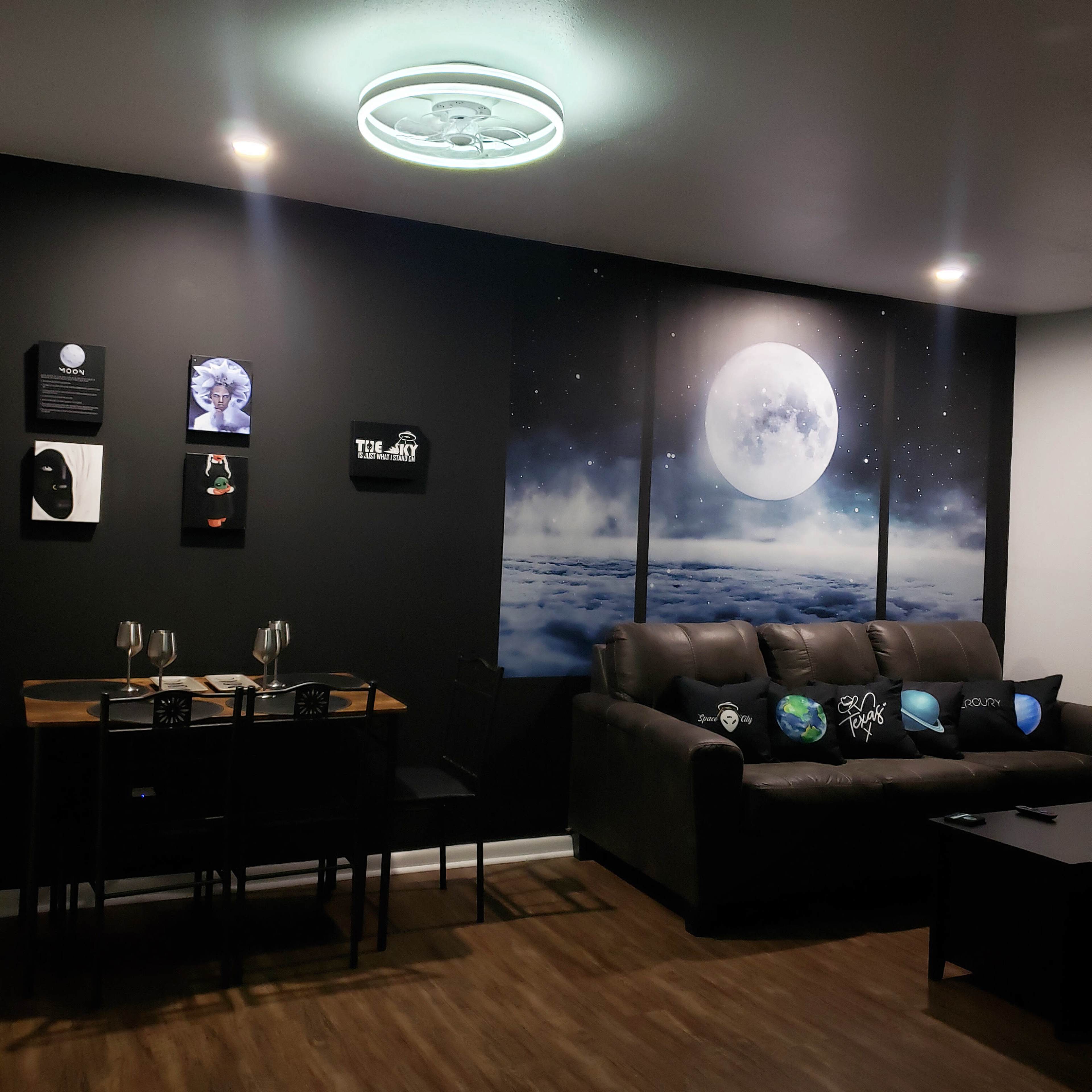 A modern living room with a dark wall, a large wall mural of the moon above clouds, a gray couch, a small dining table with two chairs, and various decorative pillows featuring celestial designs.