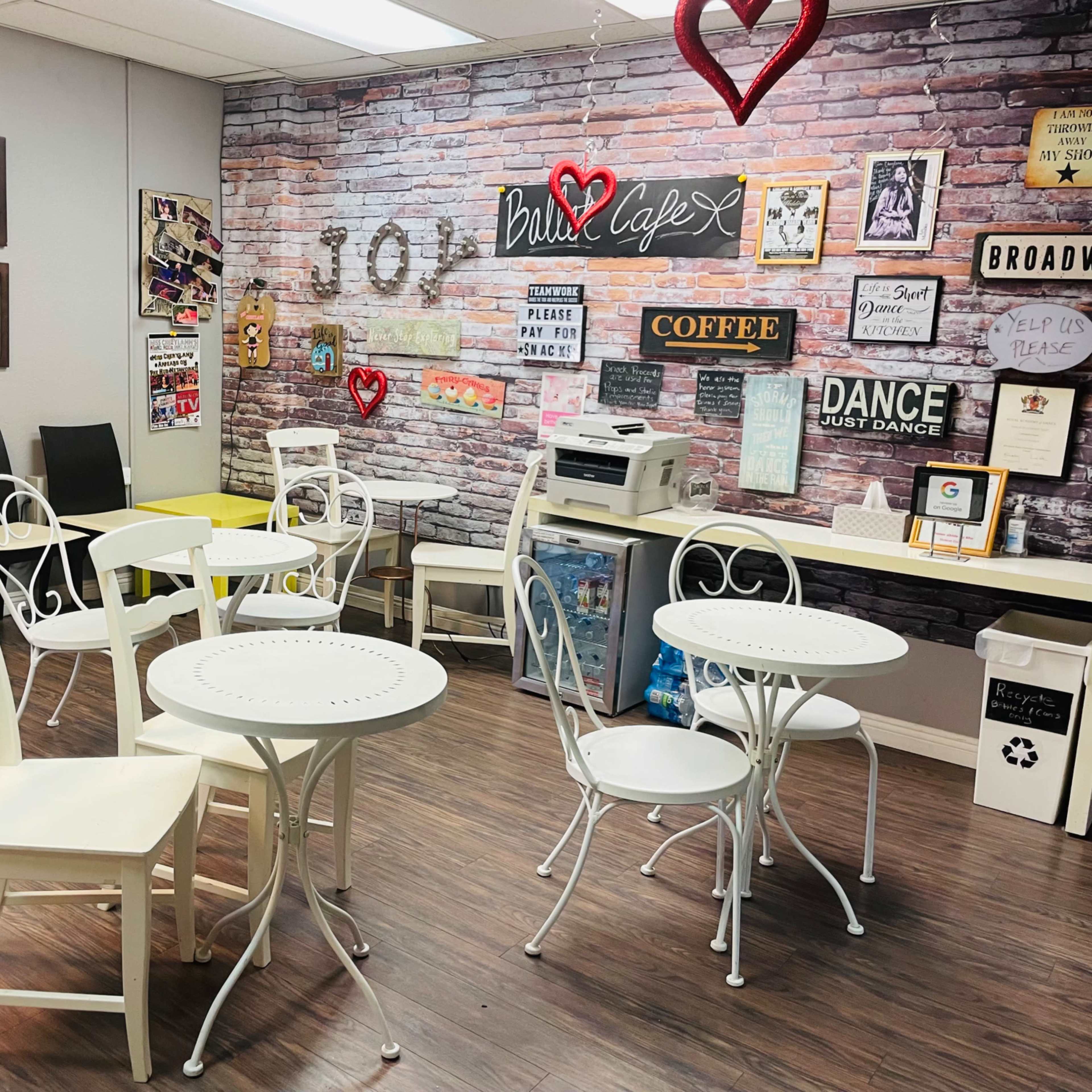 A quaint café interior with white metal tables and chairs, a printer, and a wall decorated with posters and a brick design.
