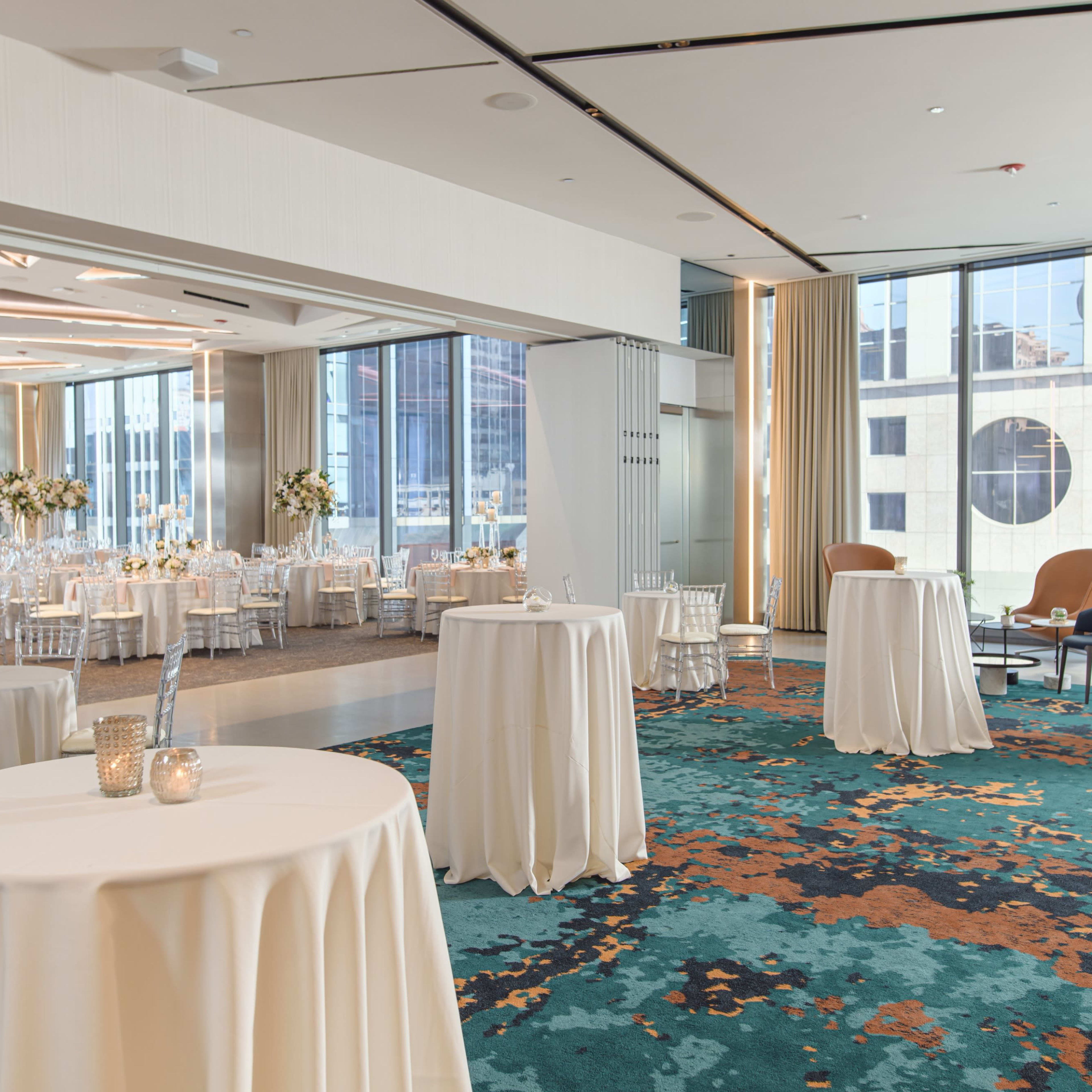 A spacious event venue with round tables covered in white tablecloths, elegant seating, and large windows showcasing a city view.