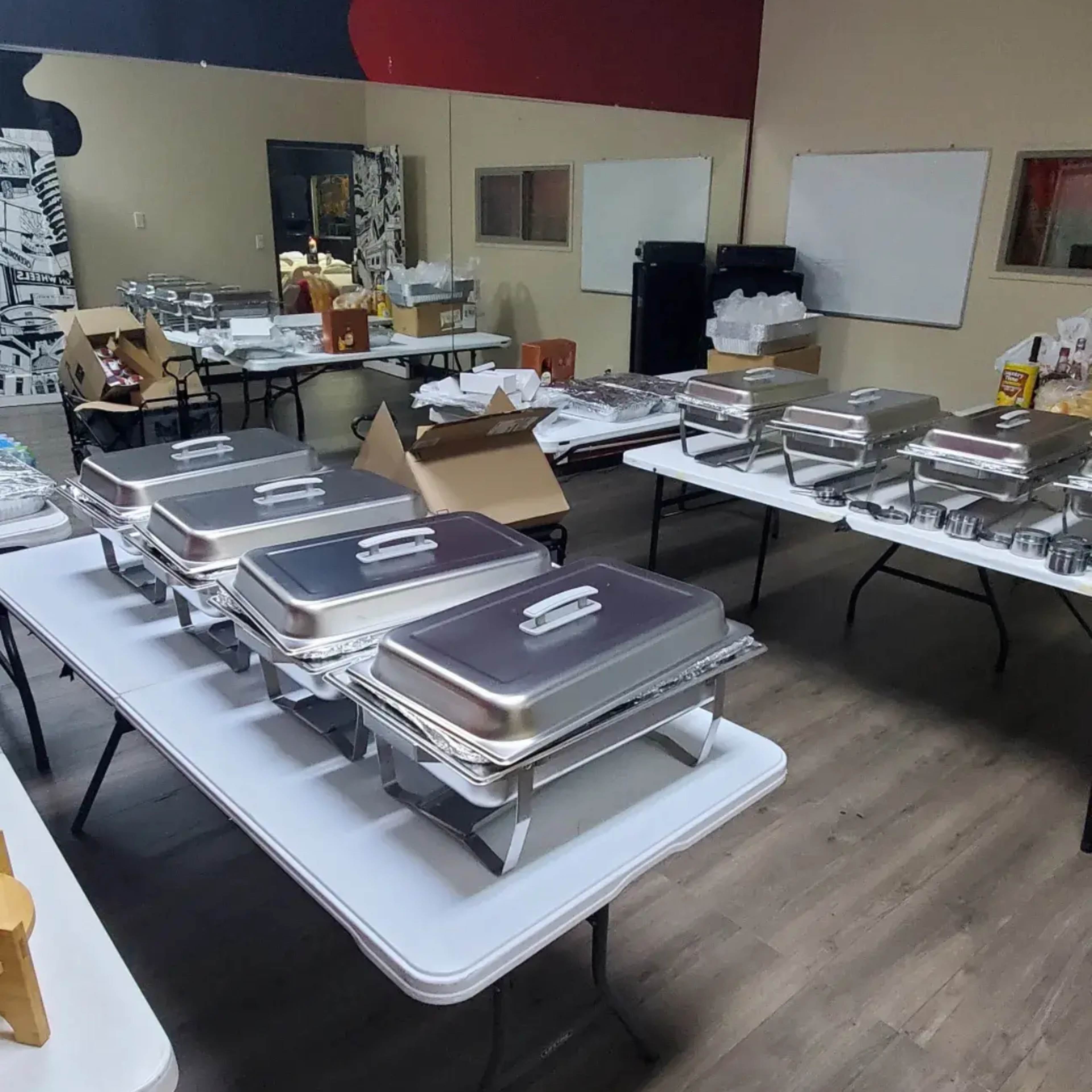 A spacious room is filled with several tables set up with chafing dishes, food containers, and scattered supplies.