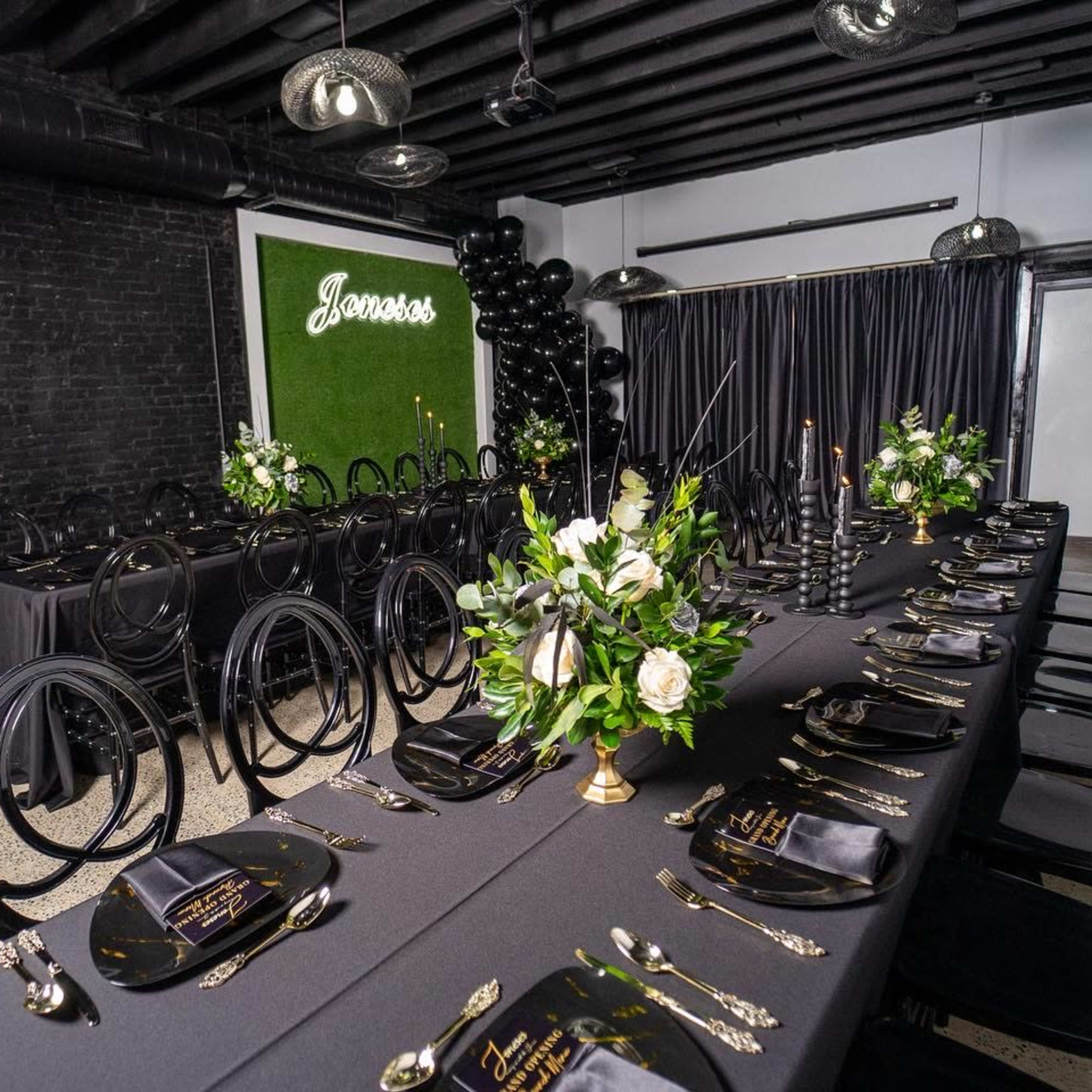 A long, elegantly set dining table with black tablecloths, black chairs, and floral centerpieces is arranged in a decorated room featuring black walls and greenery accents.