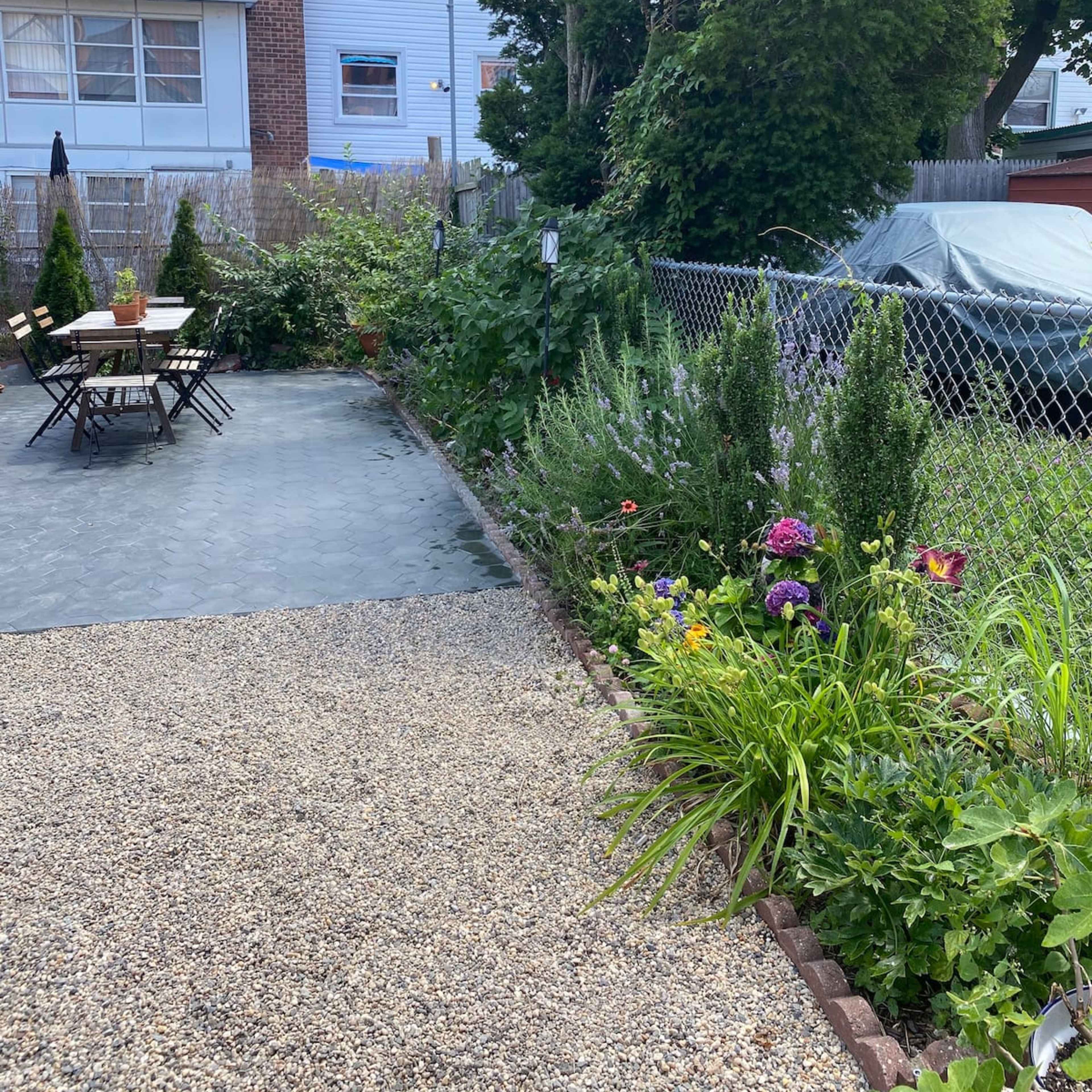 A backyard with a stone pathway leading to a table and chairs, surrounded by greenery and flowers, with a car partially covered in a tarp nearby.