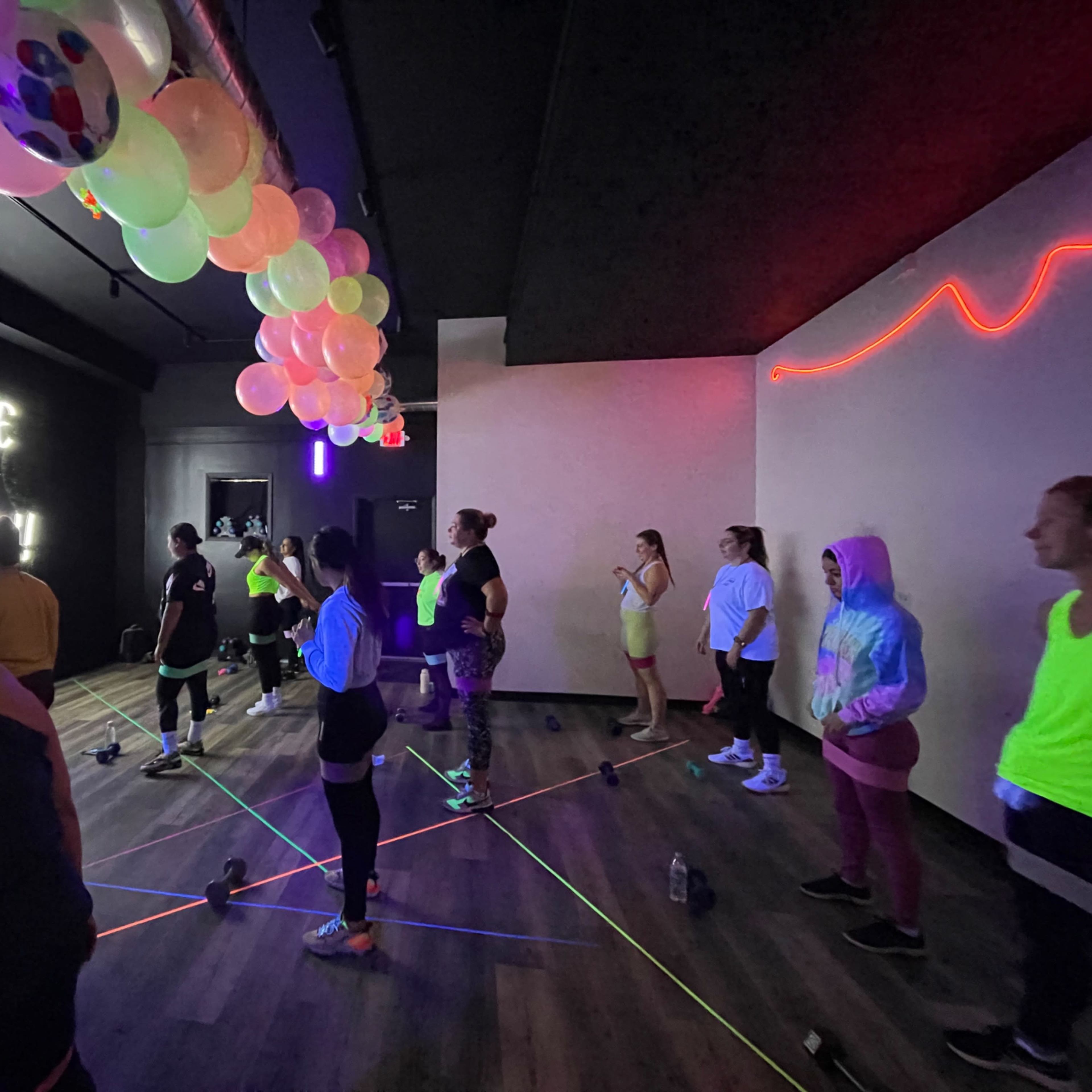 A group of people participate in a vibrant fitness class in a gym decorated with colorful balloons and neon lights.