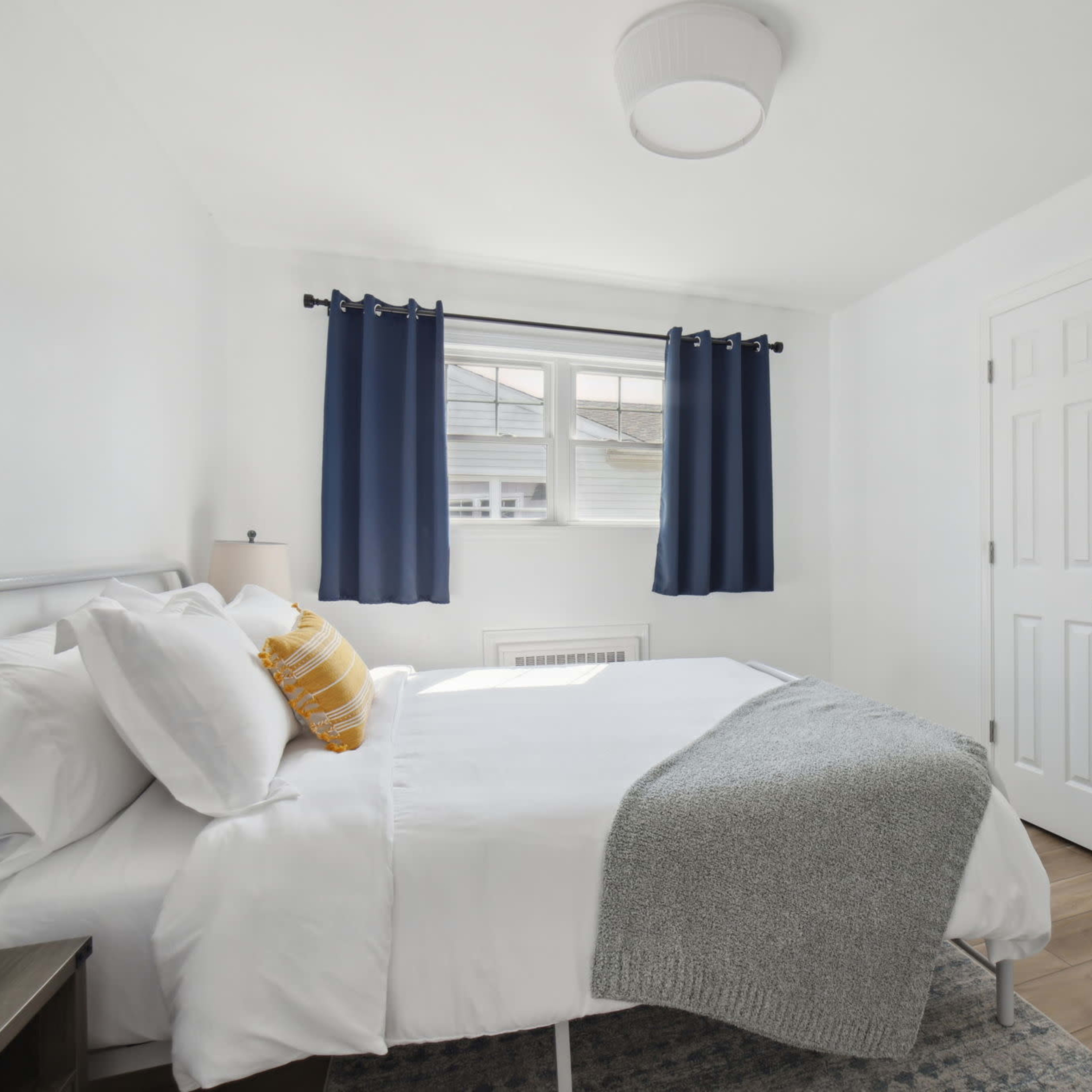 A small, neatly arranged bedroom featuring a bed with white linens, a gray throw blanket, and blue curtains over a window.