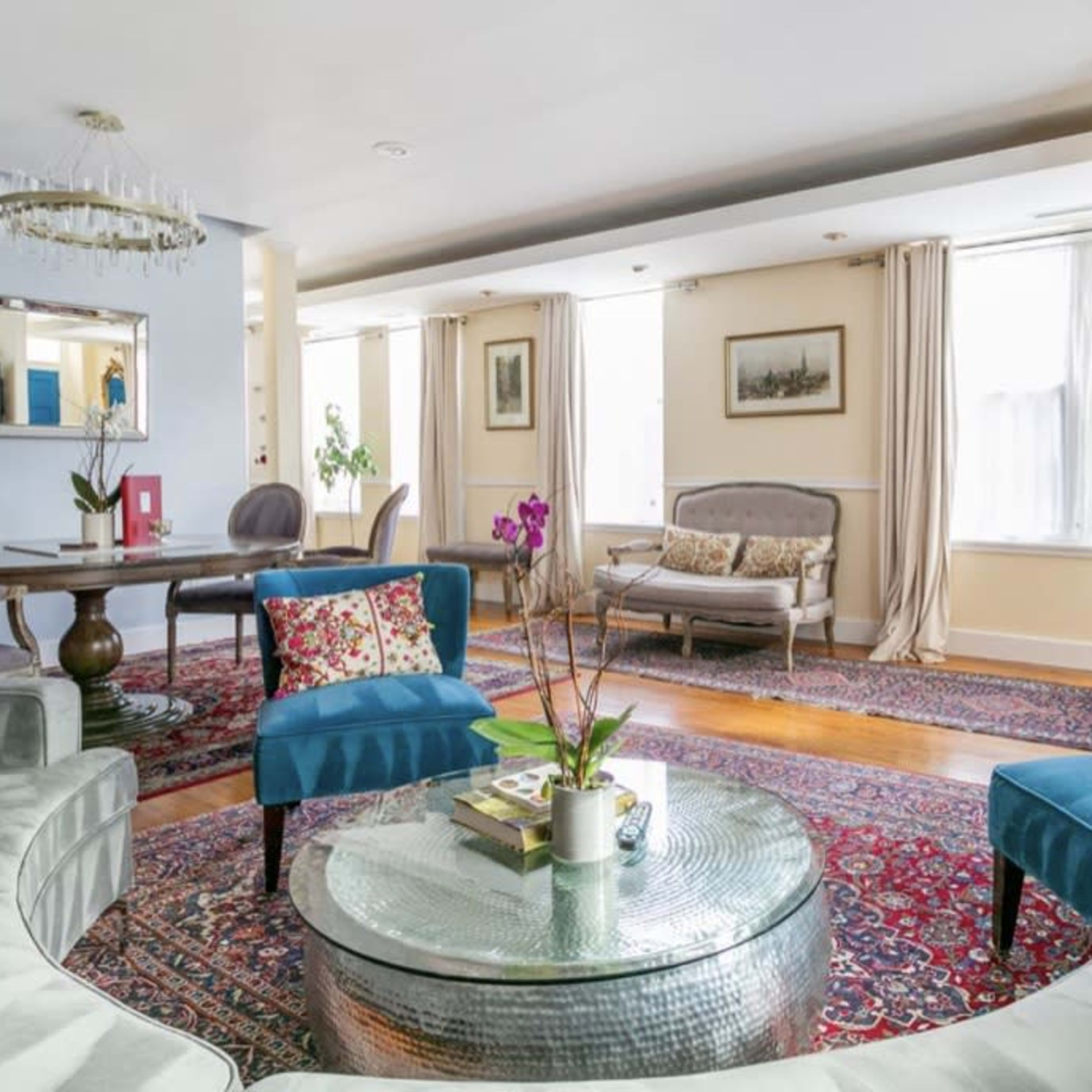 A modern living room with a curved gray sofa, blue accent chairs, a glass coffee table, and a dining area in the background, featuring a chandelier and decorative artwork on the walls.