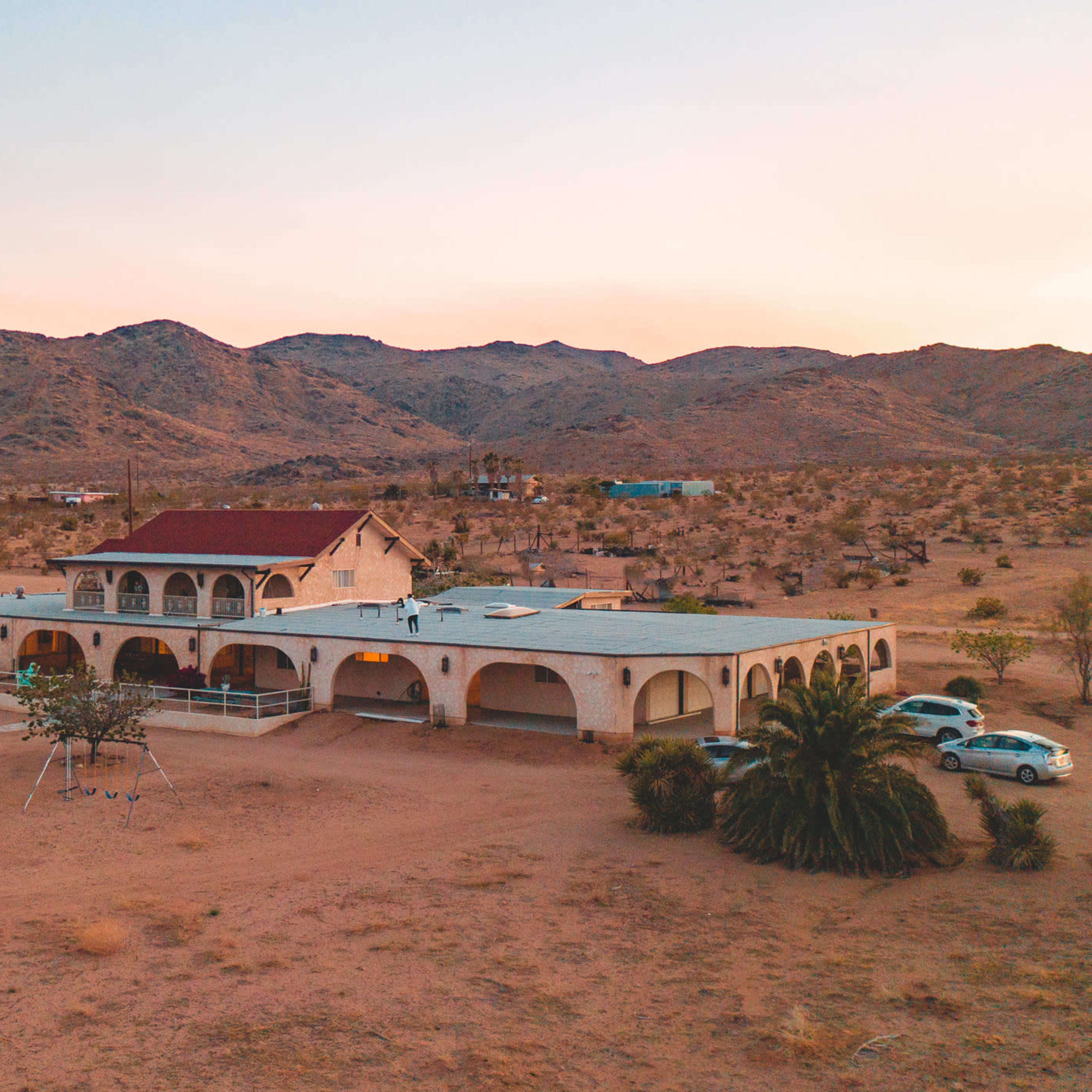 A large, multi-level house with arched features situated in a desert landscape surrounded by mountains.