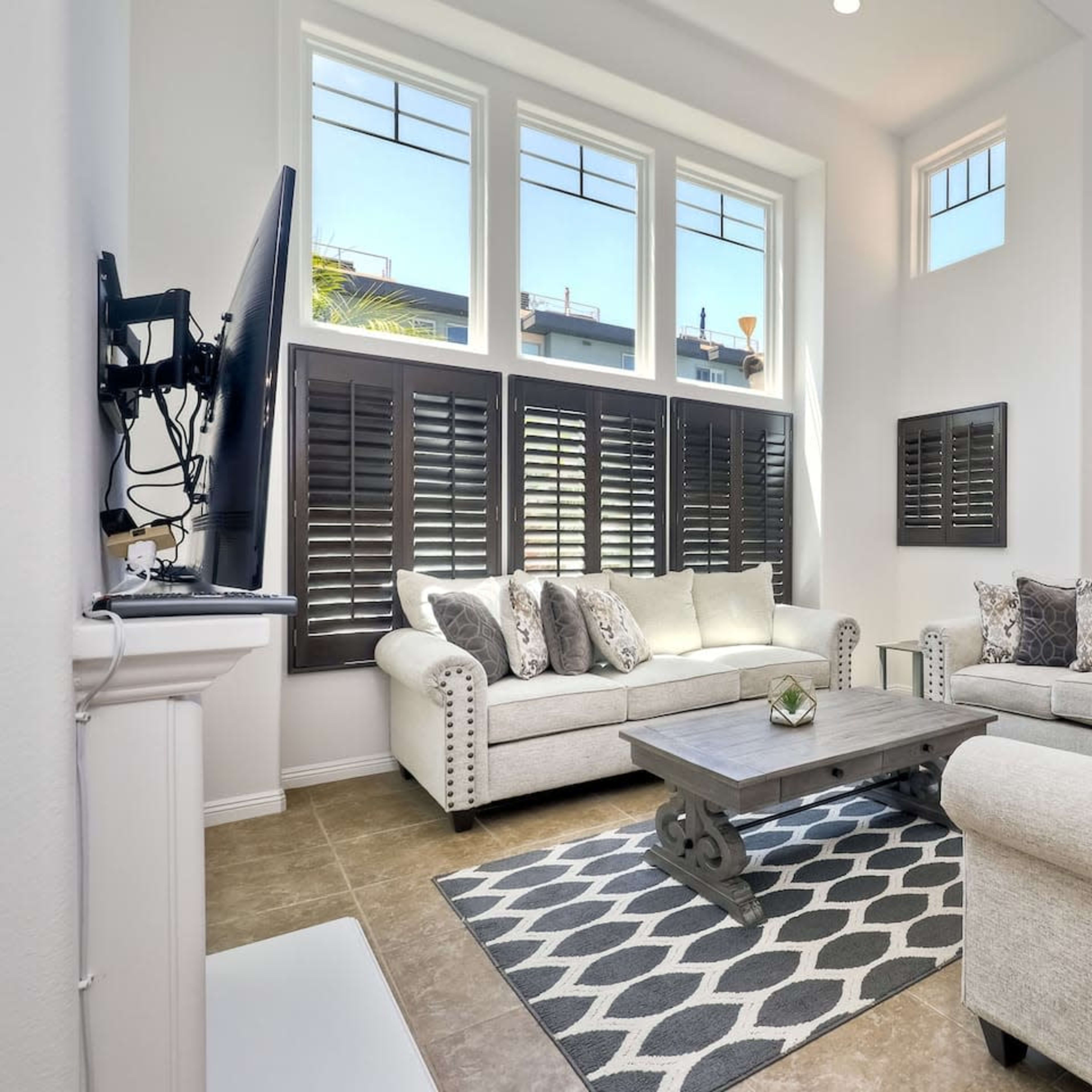 A spacious living room with large windows, light-colored furniture, and a coffee table on a patterned rug.
