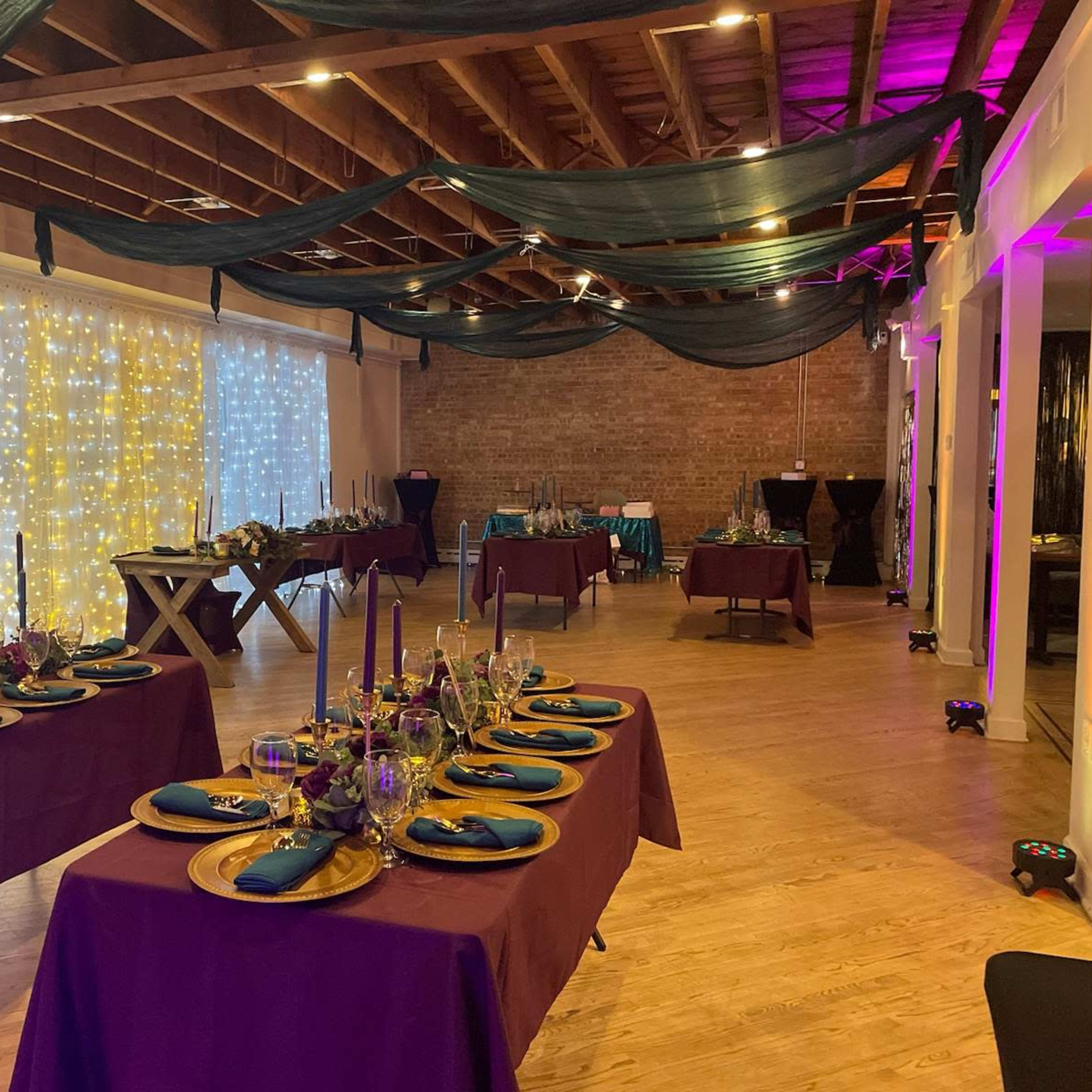 A banquet hall decorated with dim lighting, featuring tables set with blue napkins, gold plates, and floral centerpieces, against a backdrop of twinkling fairy lights.