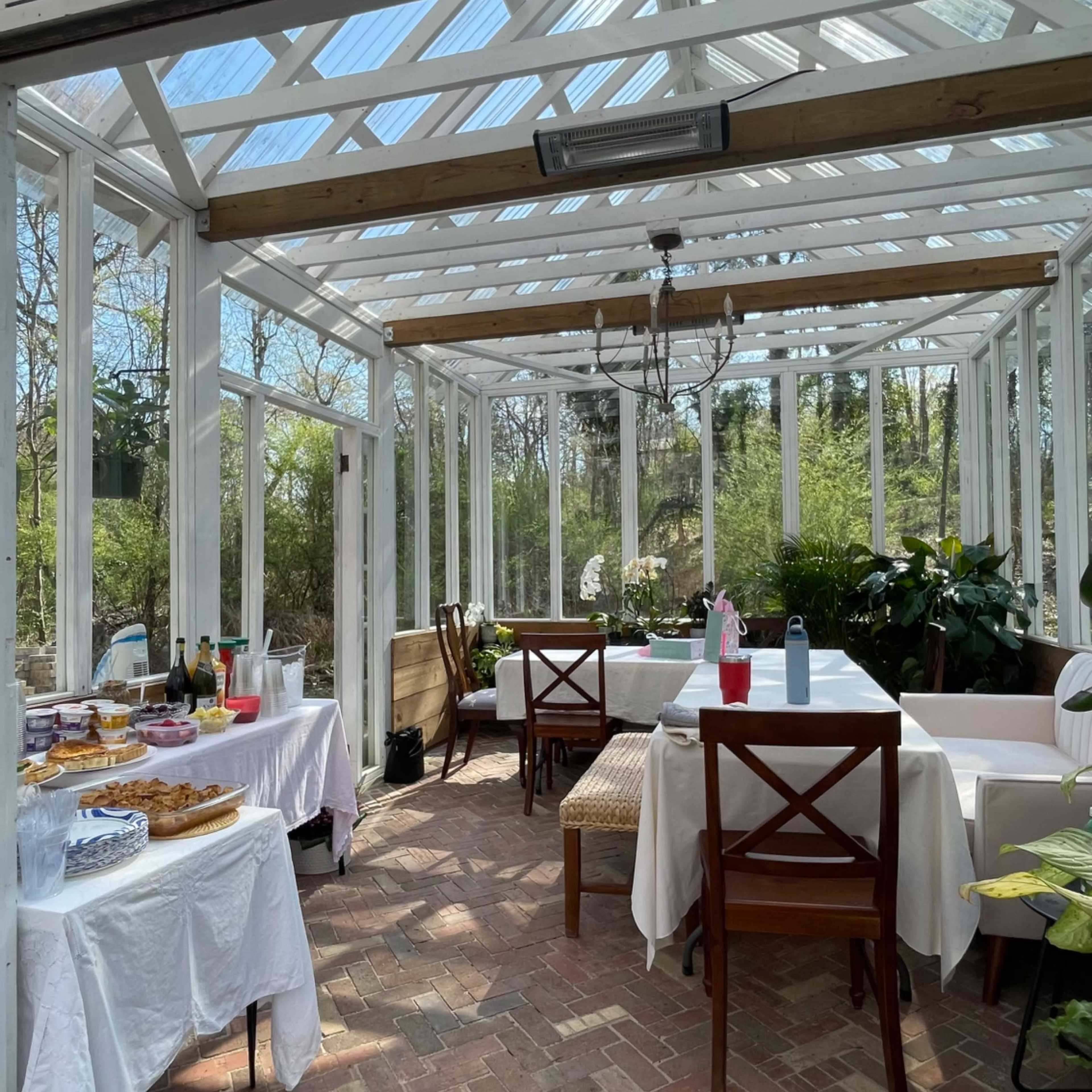 A bright, airy greenhouse with wooden tables draped in white cloths and a variety of food laid out, surrounded by greenery.