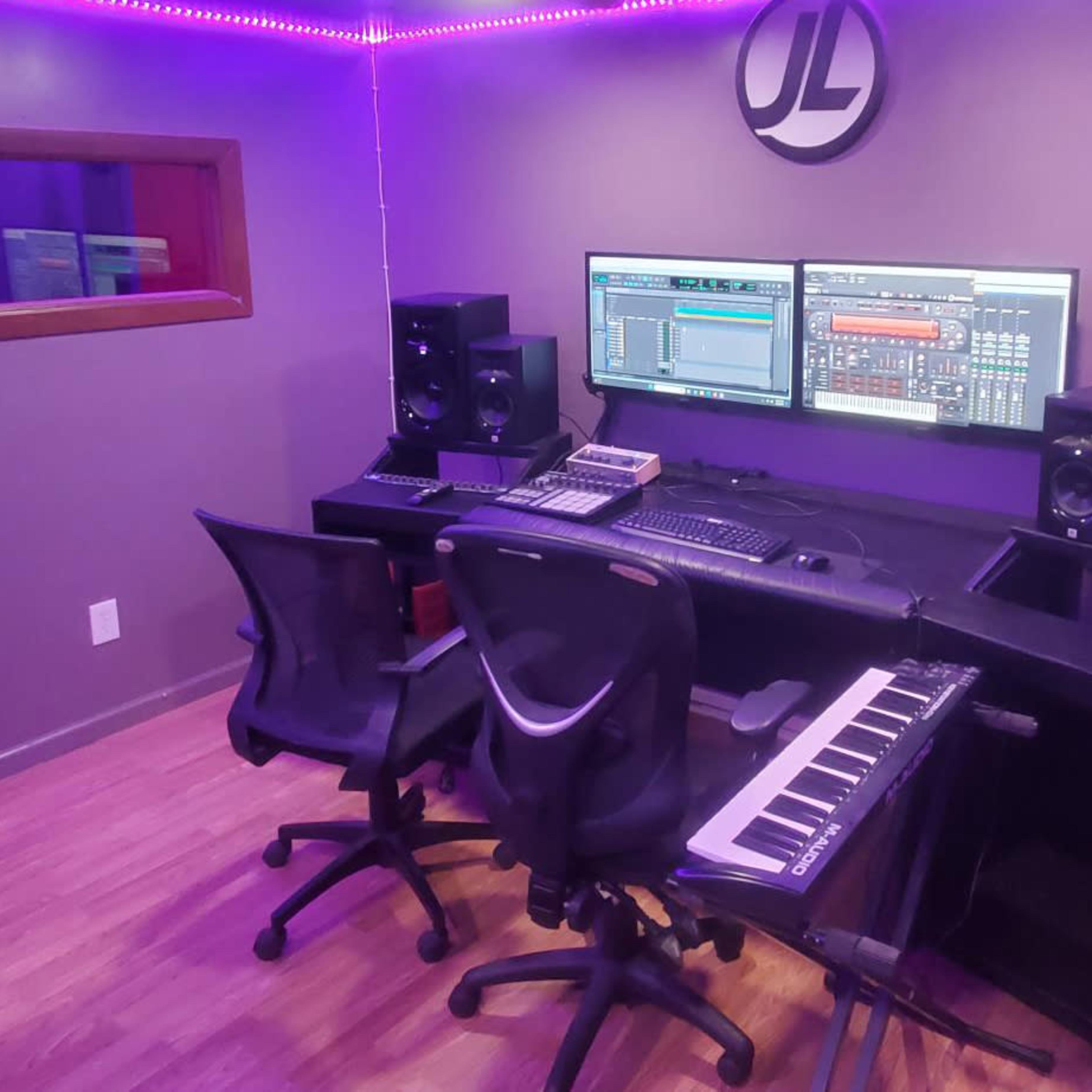 A modern music production studio featuring a dual monitor setup, audio speakers, a keyboard, and two rolling chairs.