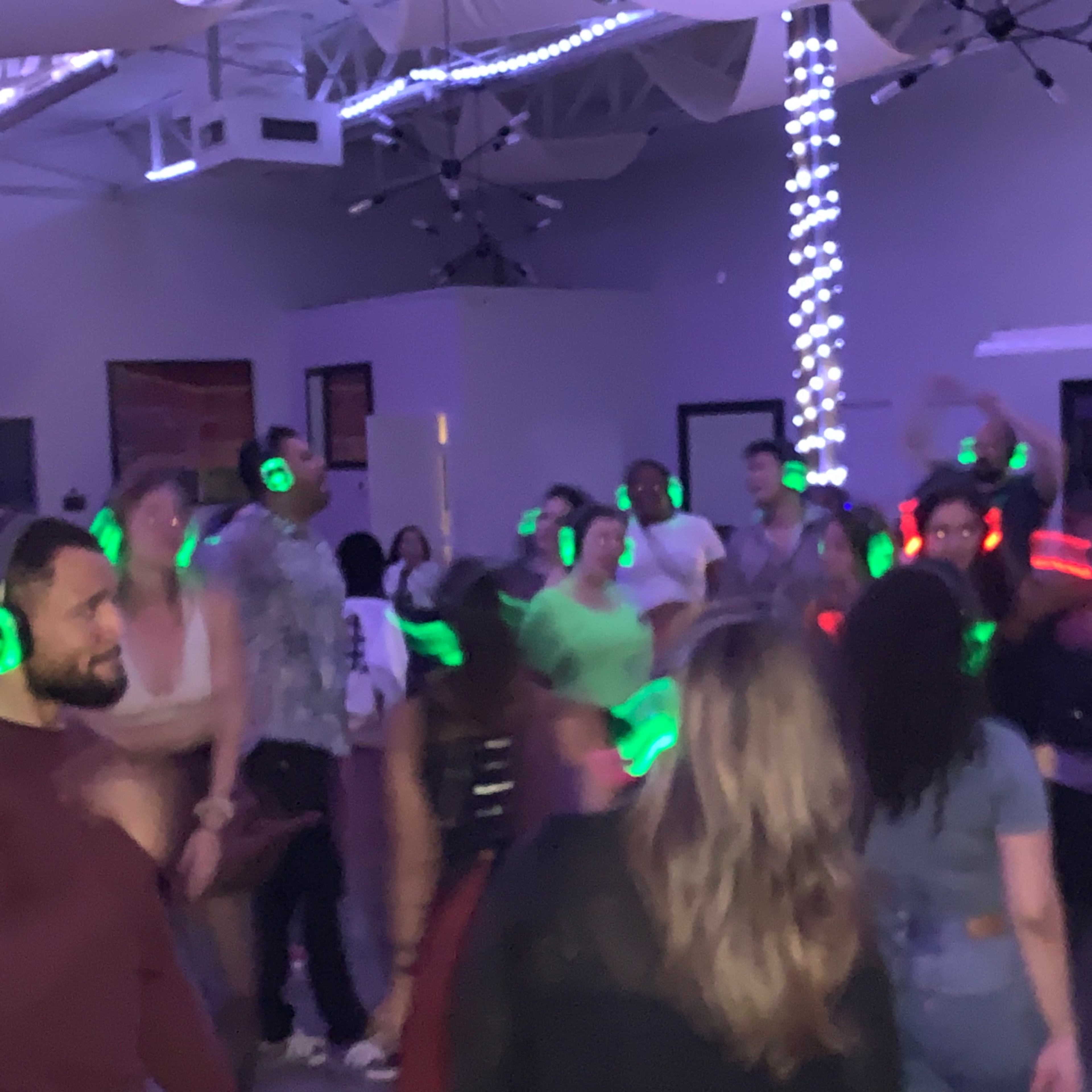 A group of people is dancing indoors at a party while wearing wireless headphones with glowing lights.