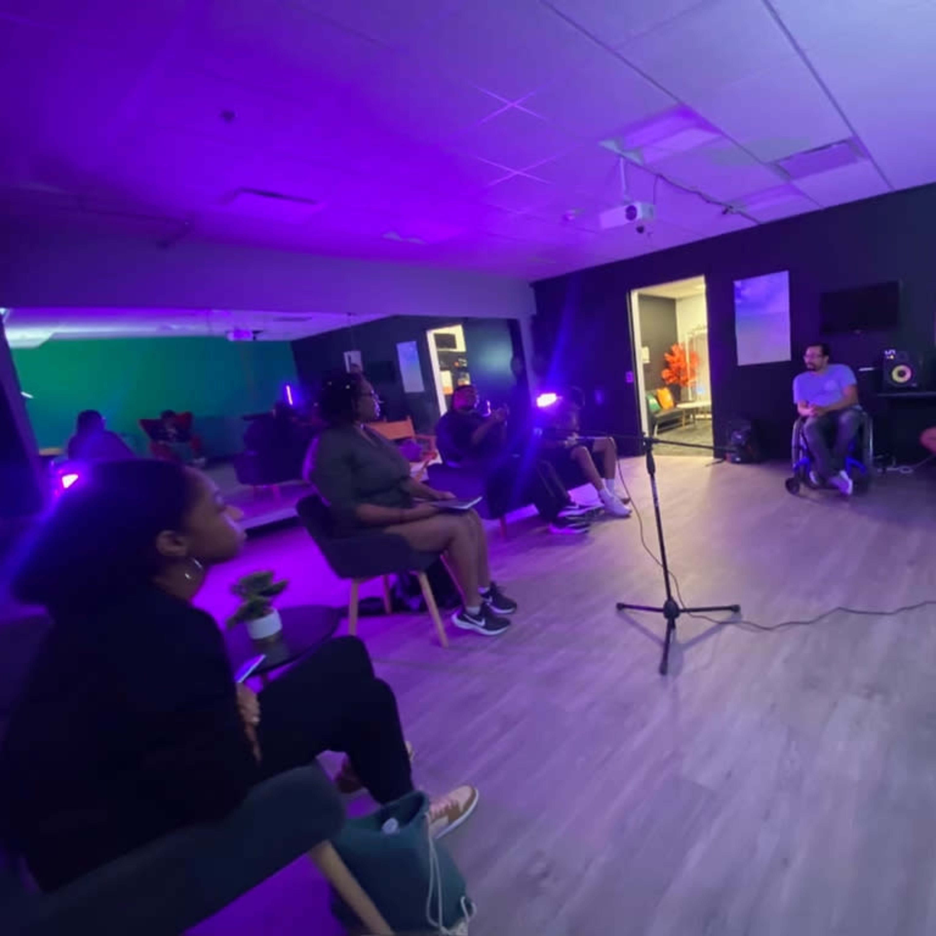 A group of people is gathered in a dimly lit room equipped with colorful lighting and technology, engaged in a discussion around a microphone.