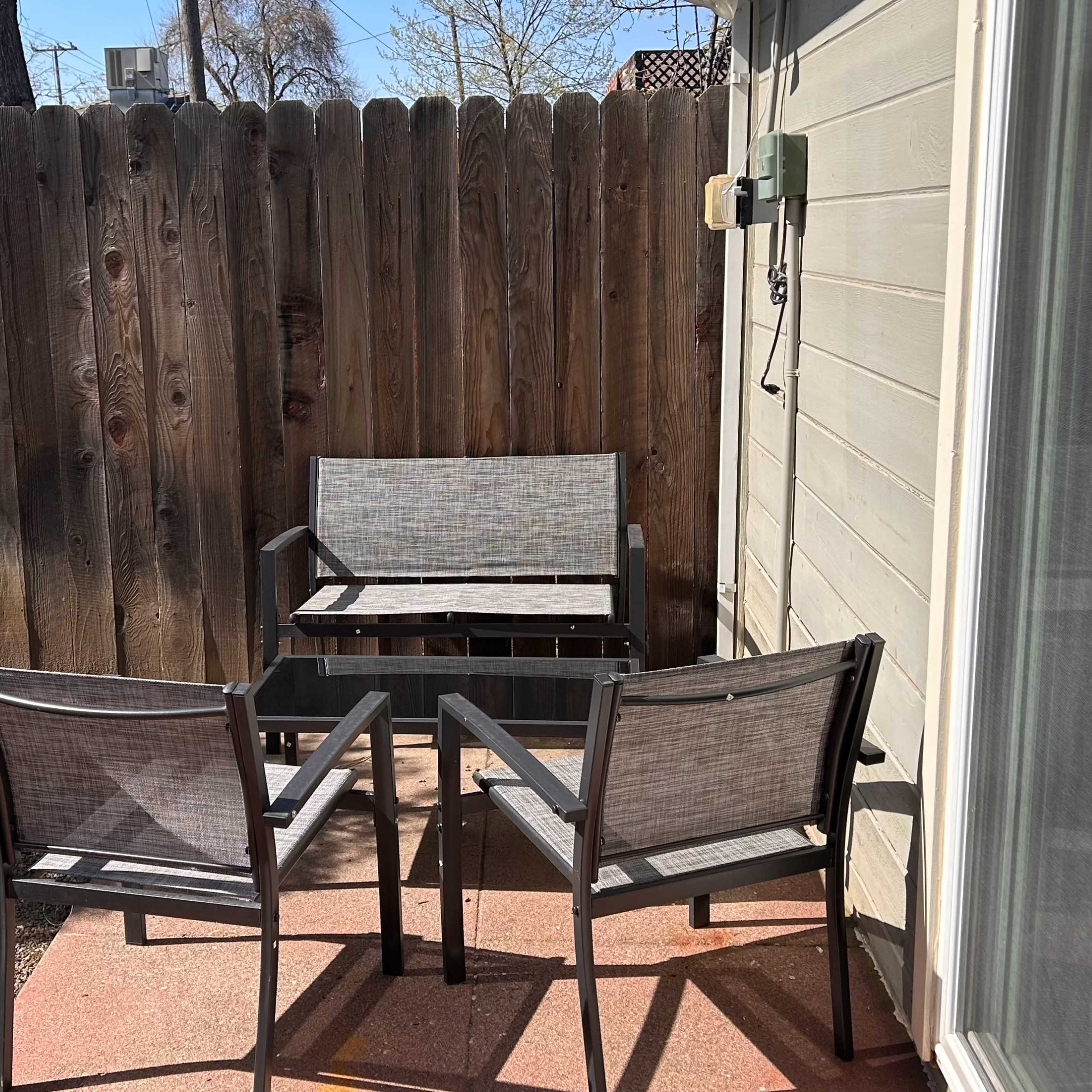 A small patio area features two chairs and a loveseat positioned against a wooden fence, surrounded by gravel and some greenery.