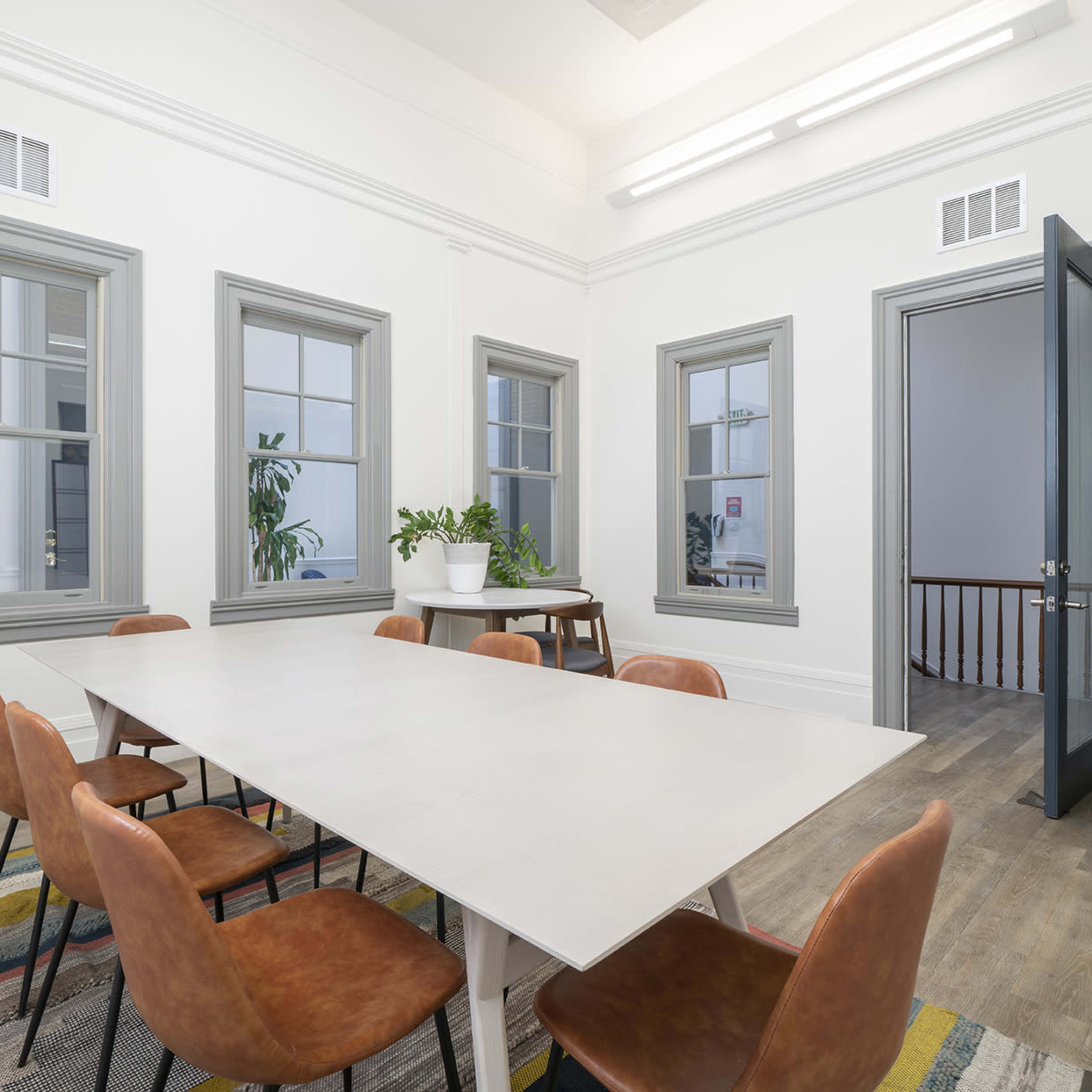 A modern meeting room features a long table surrounded by brown chairs, with large windows and light-colored walls.