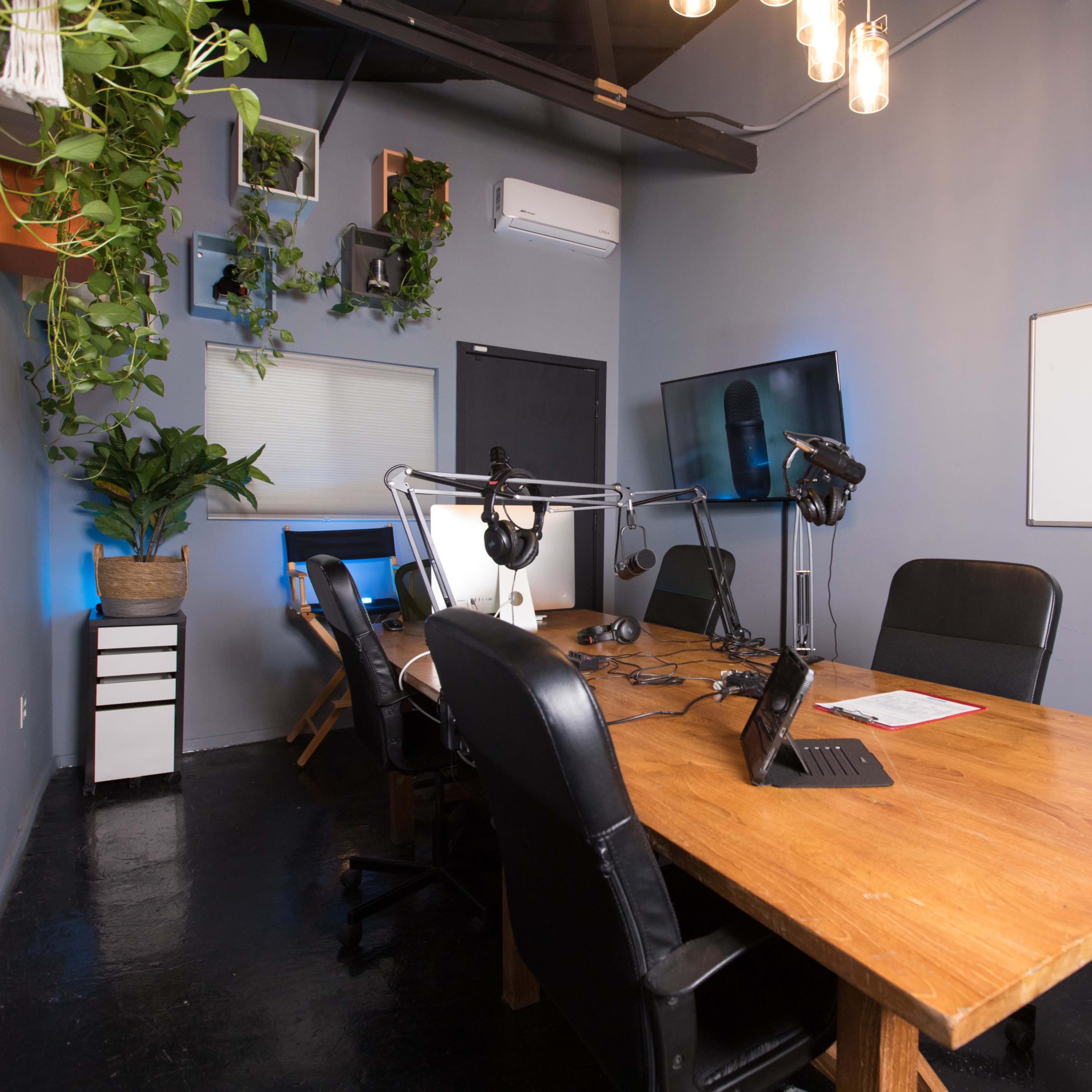 A modern conference room features a large wooden table surrounded by black chairs, equipped with microphones and computer screens, and adorned with plants and wall decorations.
