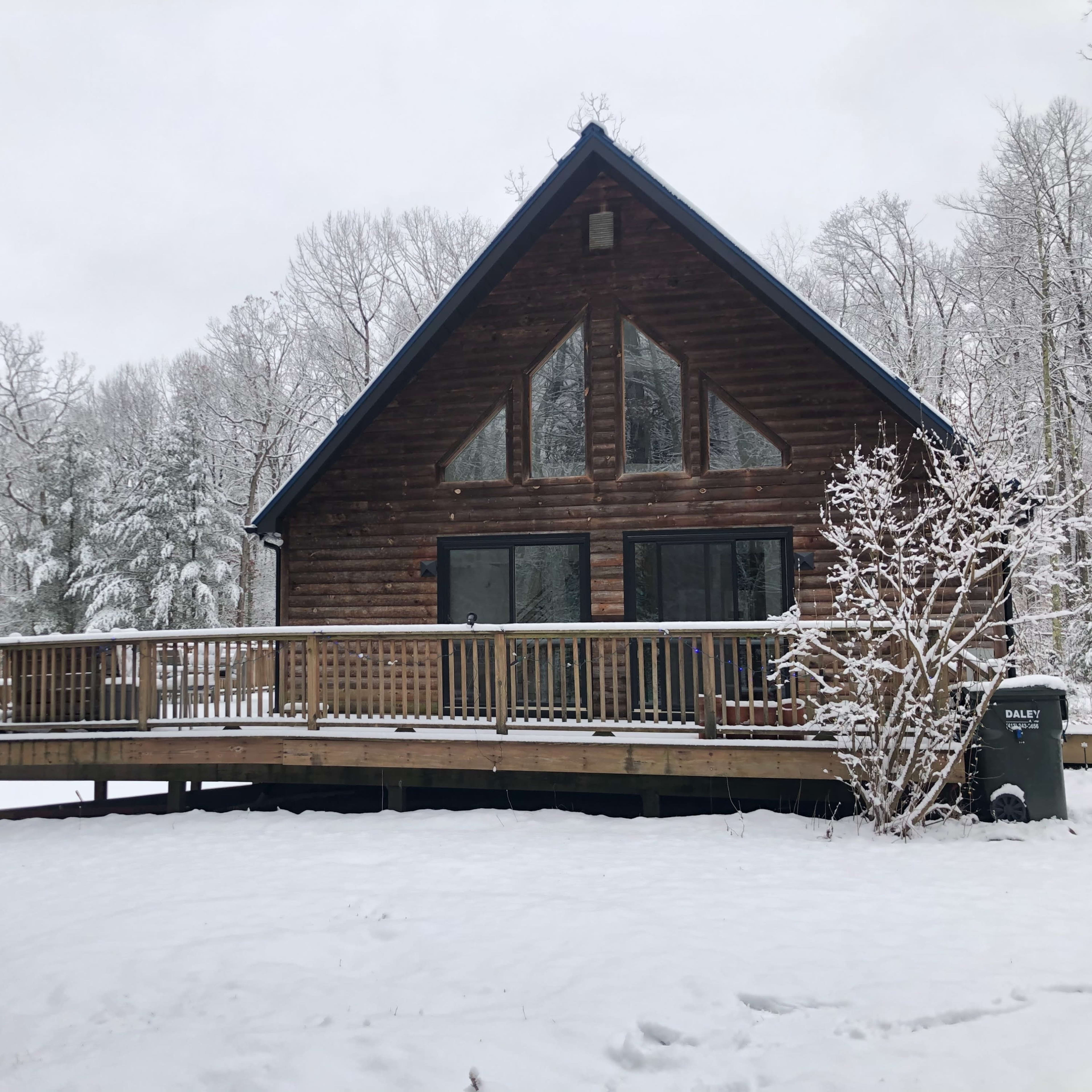 A wooden cabin with a deck is surrounded by snow-covered trees and a flagpole displaying the American flag.
