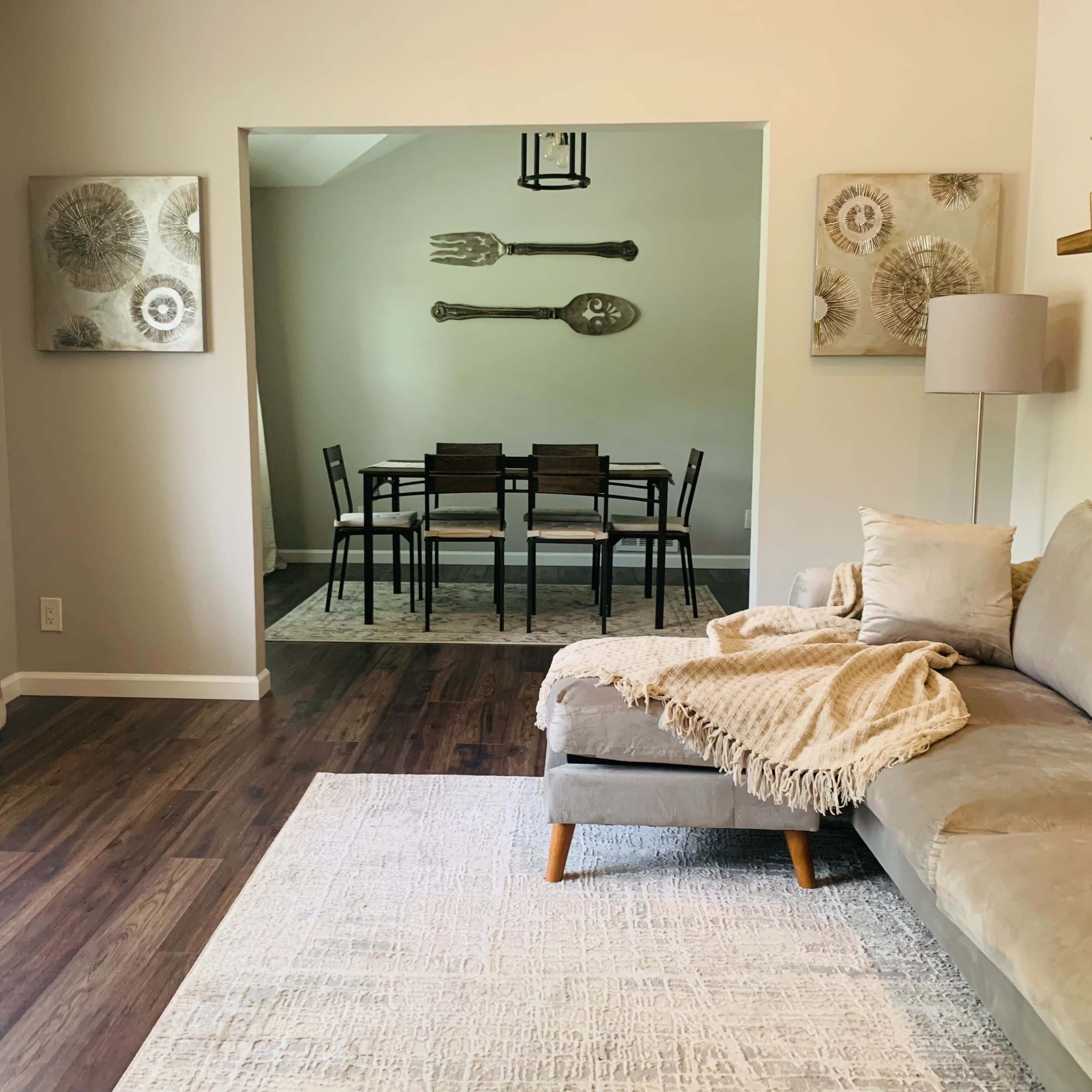 A light-filled living room features a gray sectional sofa with a blanket, leading into a dining area with a table and chairs, adorned by wall art depicting kitchen utensils.