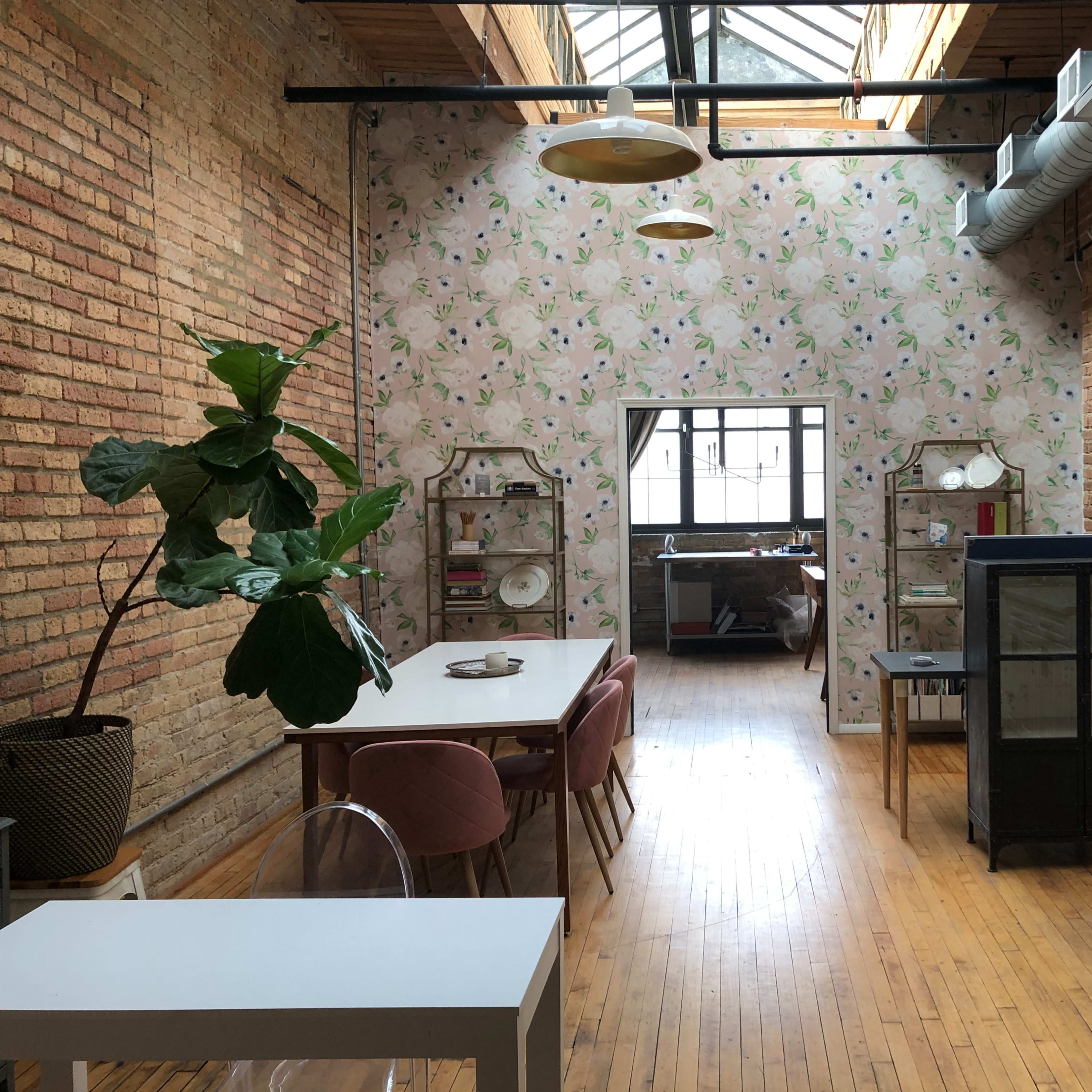 An open, well-lit interior space featuring a dining area with a white table, a decorative plant, and a view through an archway into a bedroom.