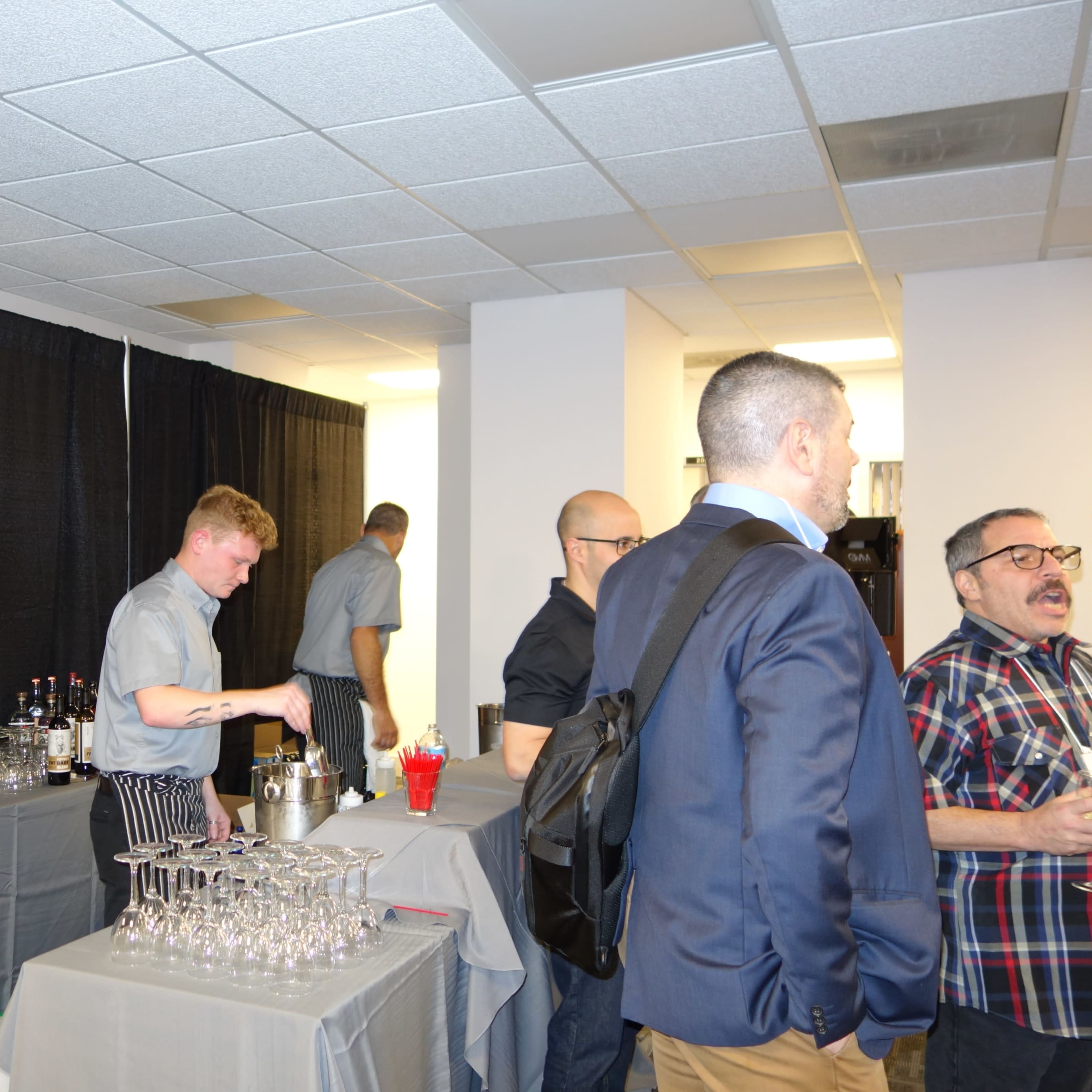 A bartender prepares drinks behind a table lined with glassware and liquor bottles at an event with attendees conversing nearby.