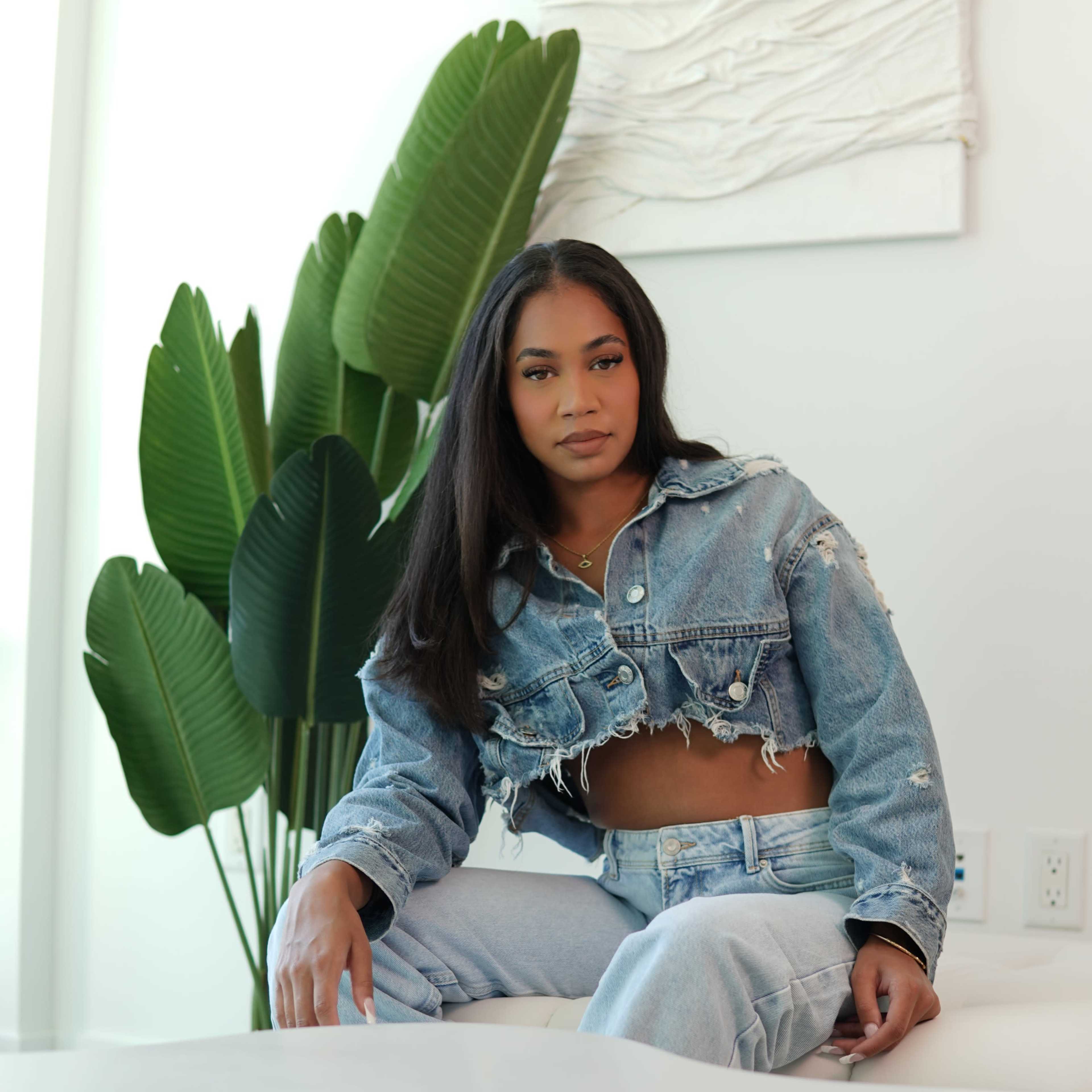 A woman sits on a white sofa next to a large leafy plant while wearing a denim cropped jacket and matching jeans.