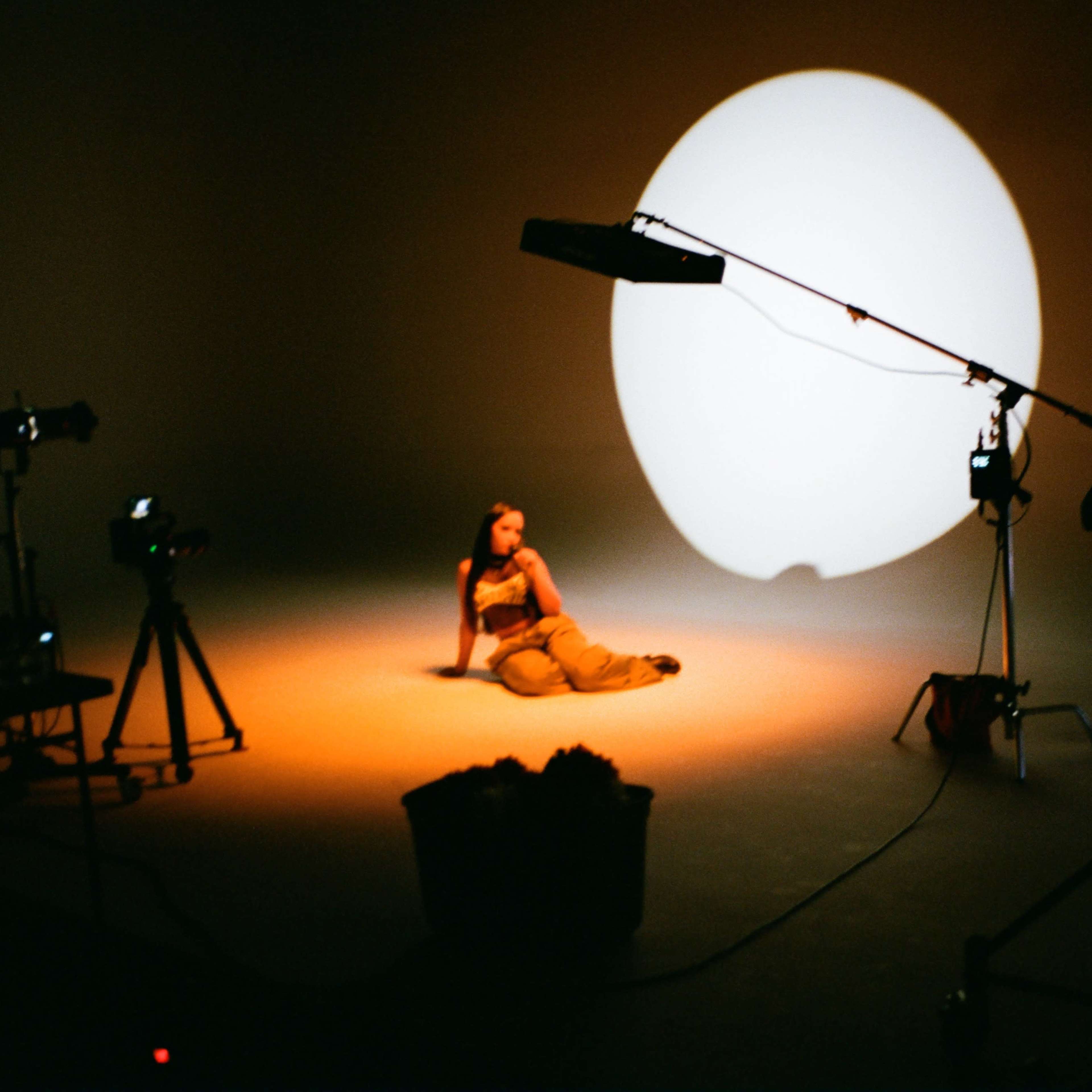 A person sits on the floor of a studio, illuminated by a large circular light in the background.