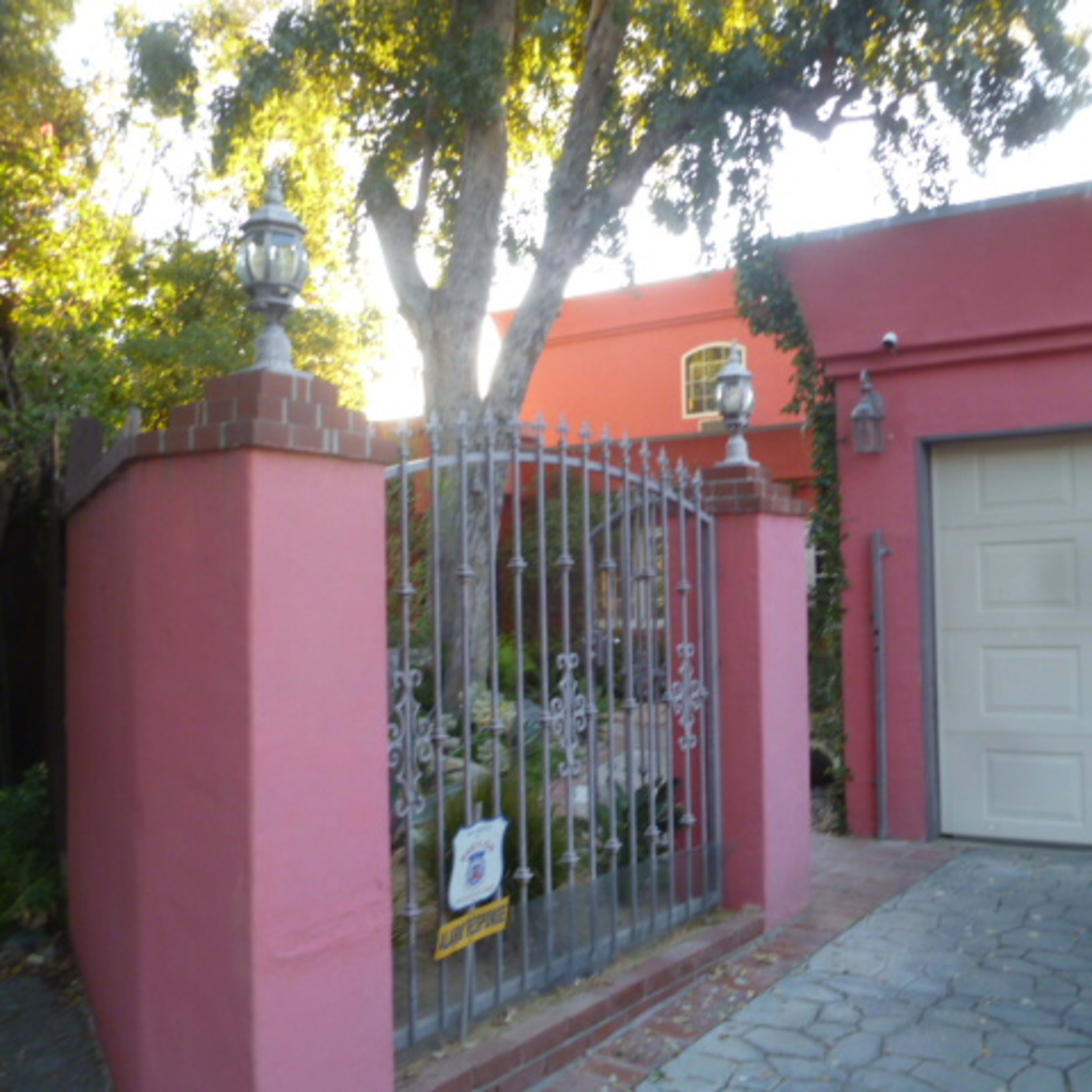 A pink stucco wall with an ornate metal gate leading to a garden and a light gray garage door beside it.